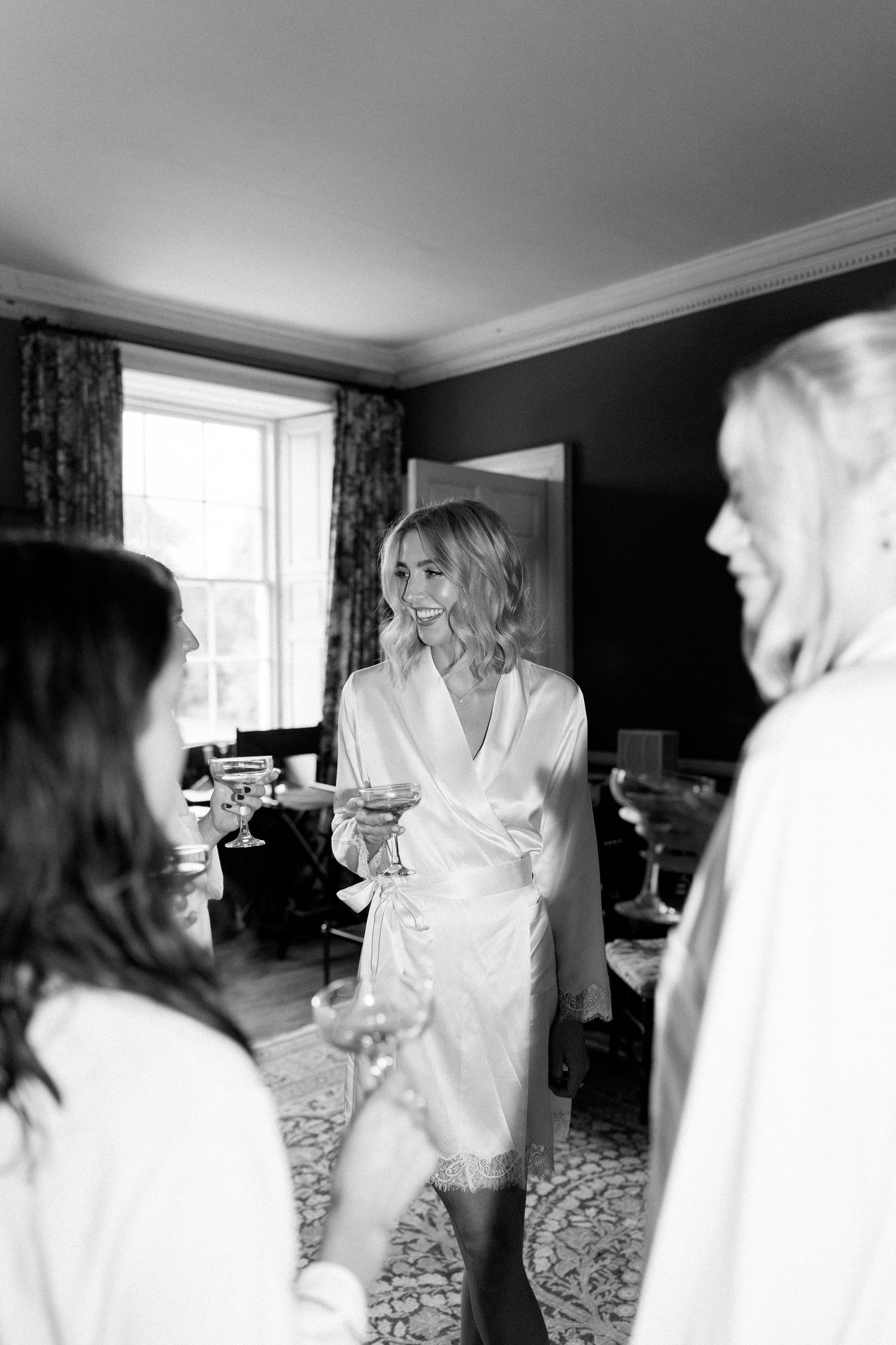 Bride in white satin robe holding champagne glass with bridesmaids during wedding morning preparations.