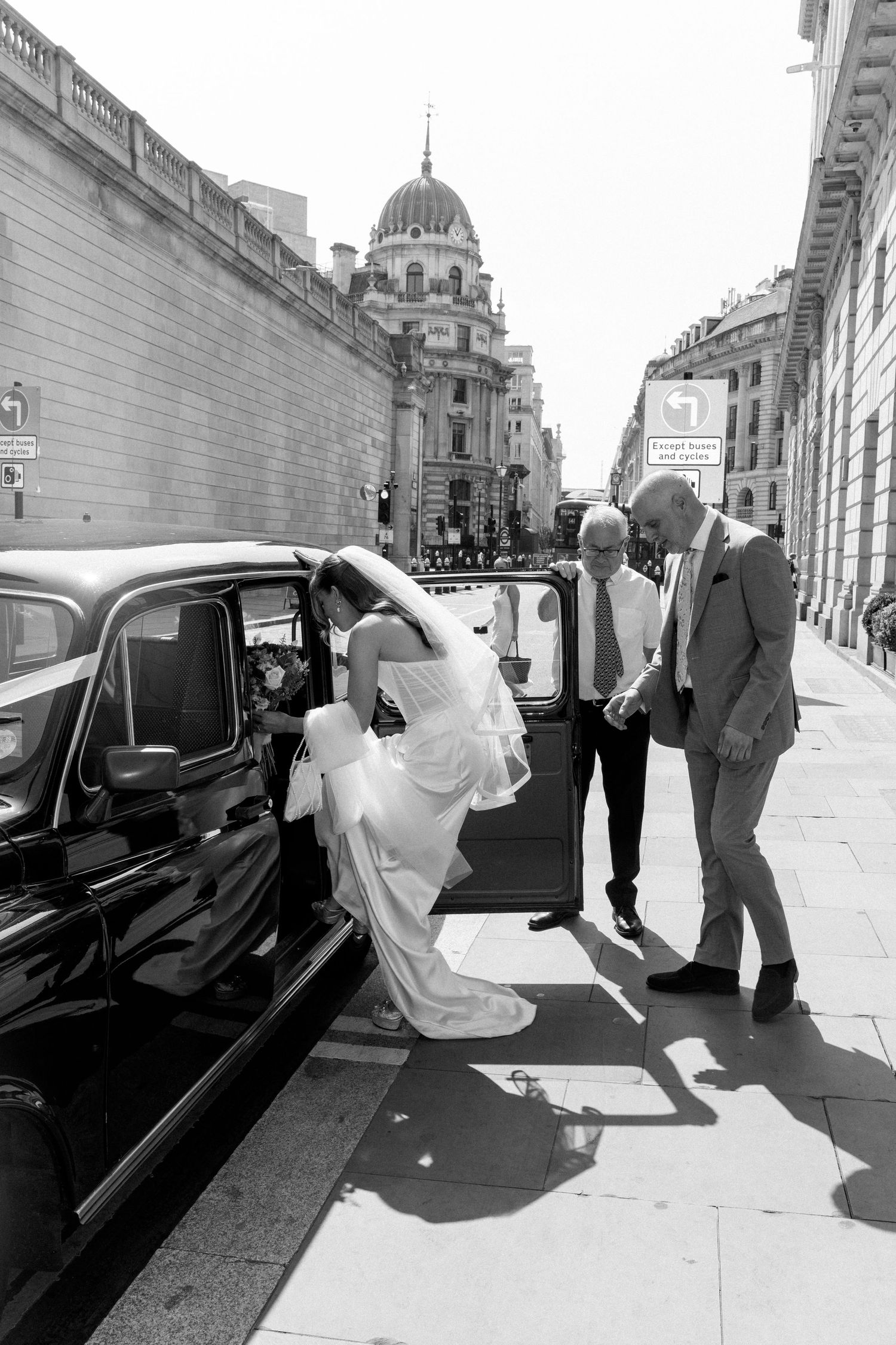Black and white photo of bride stepping out of black London taxi cab assisted by two men.
