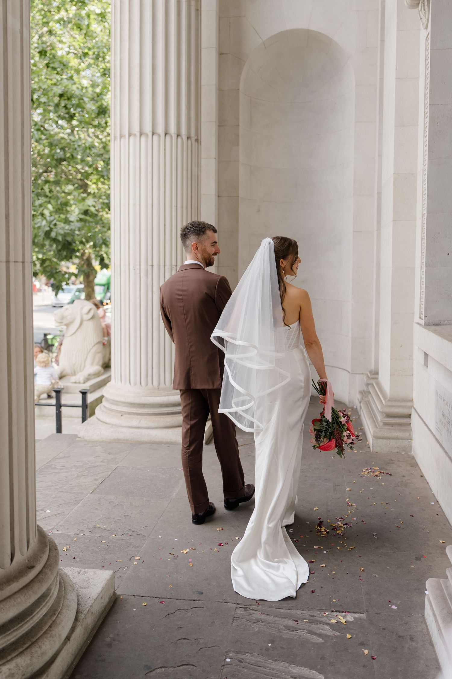 Bride in white satin gown with veil and groom in brown suit walk among classical white columns.
