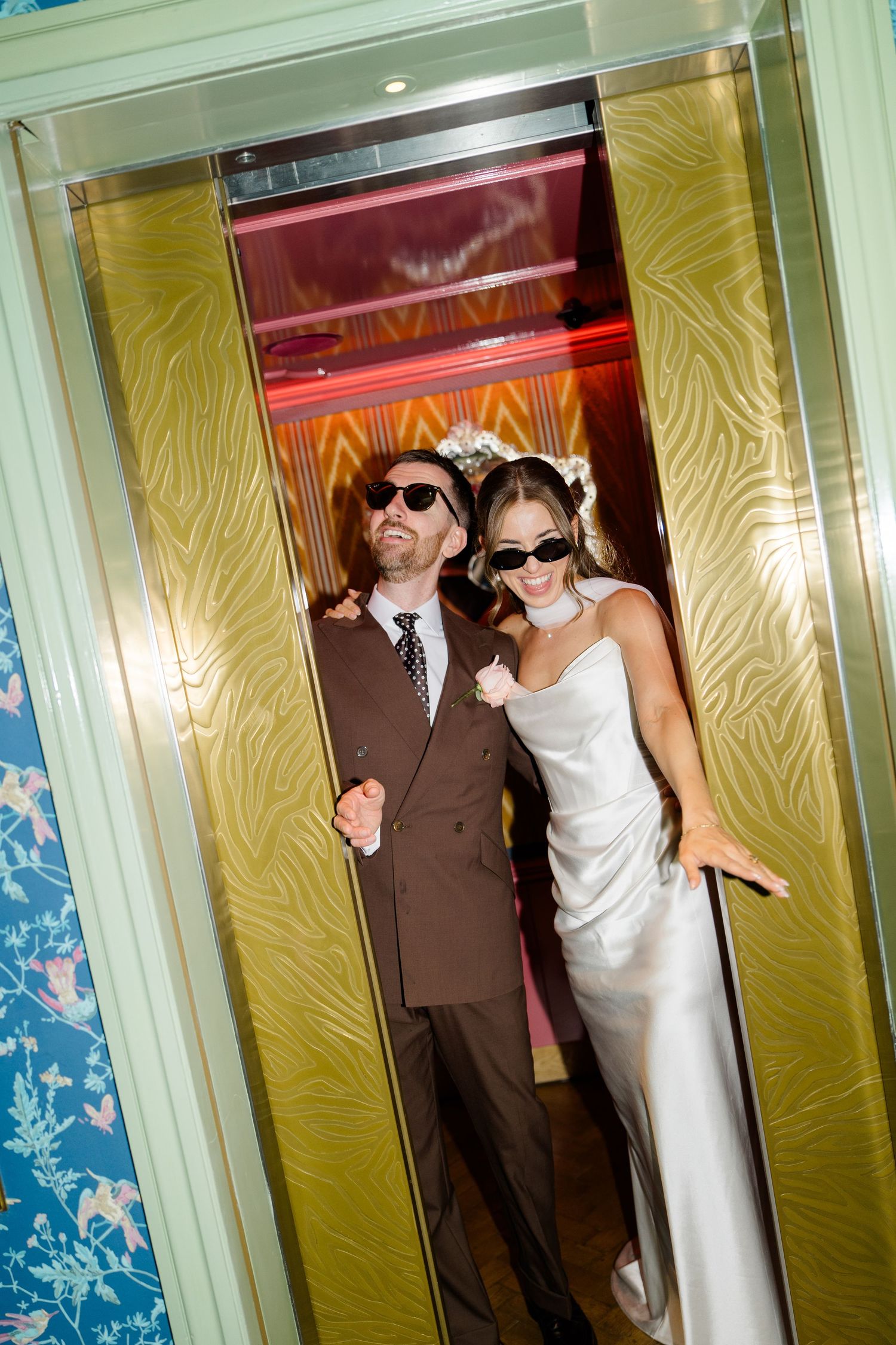 Bride and groom in sunglasses pose together stepping out of a gold Art Deco elevator.