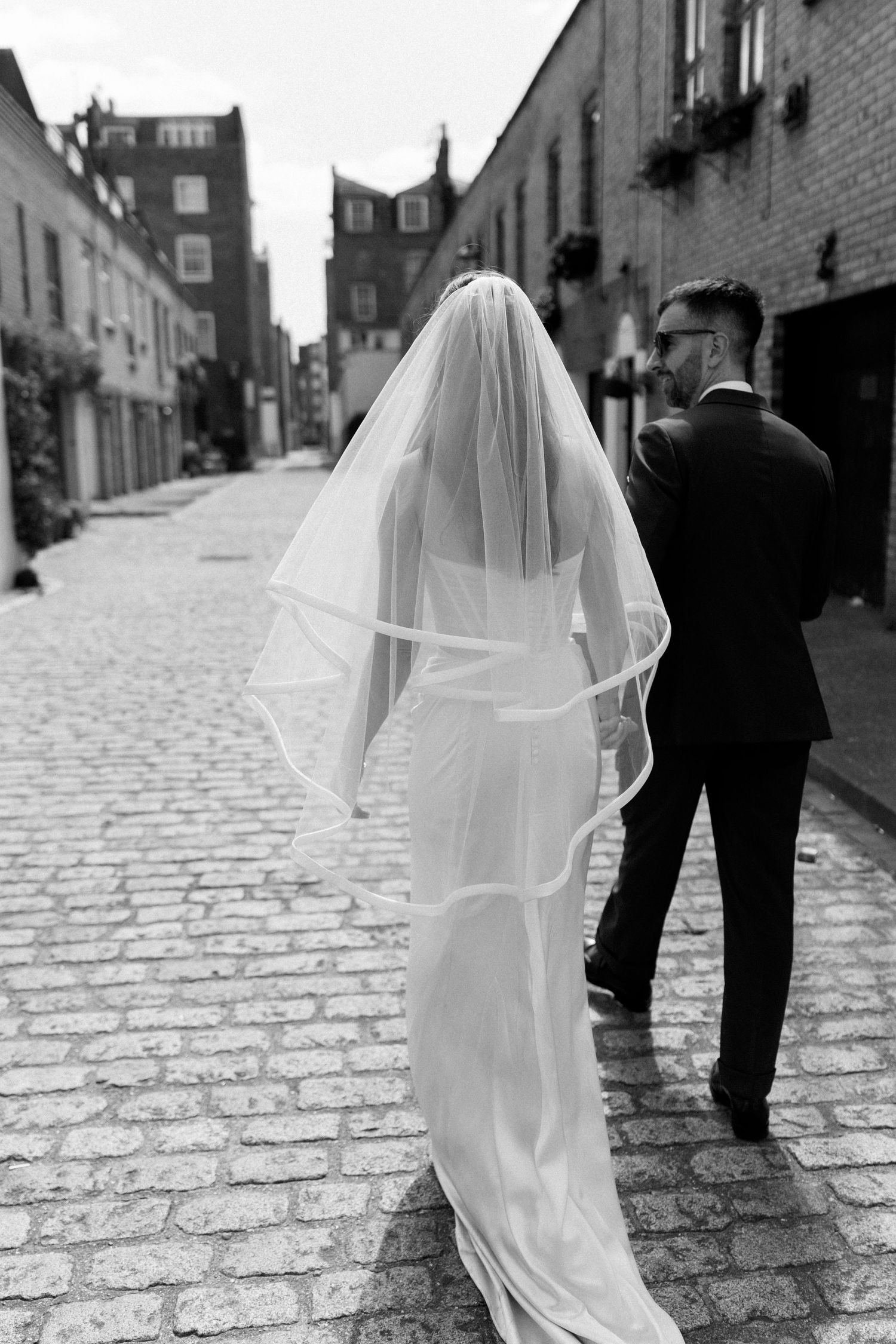 Bride in flowing veil and groom in black suit walk cobblestone street in black and white wedding photo.