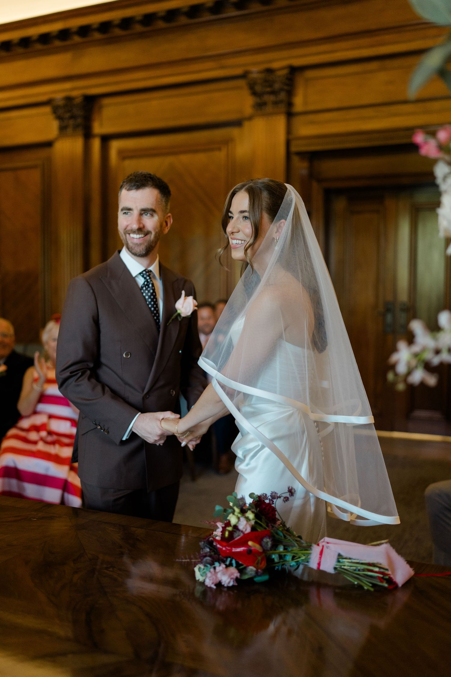 Smiling bride and groom hold hands at wedding ceremony altar, bride wearing white gown and veil with red bouquet.