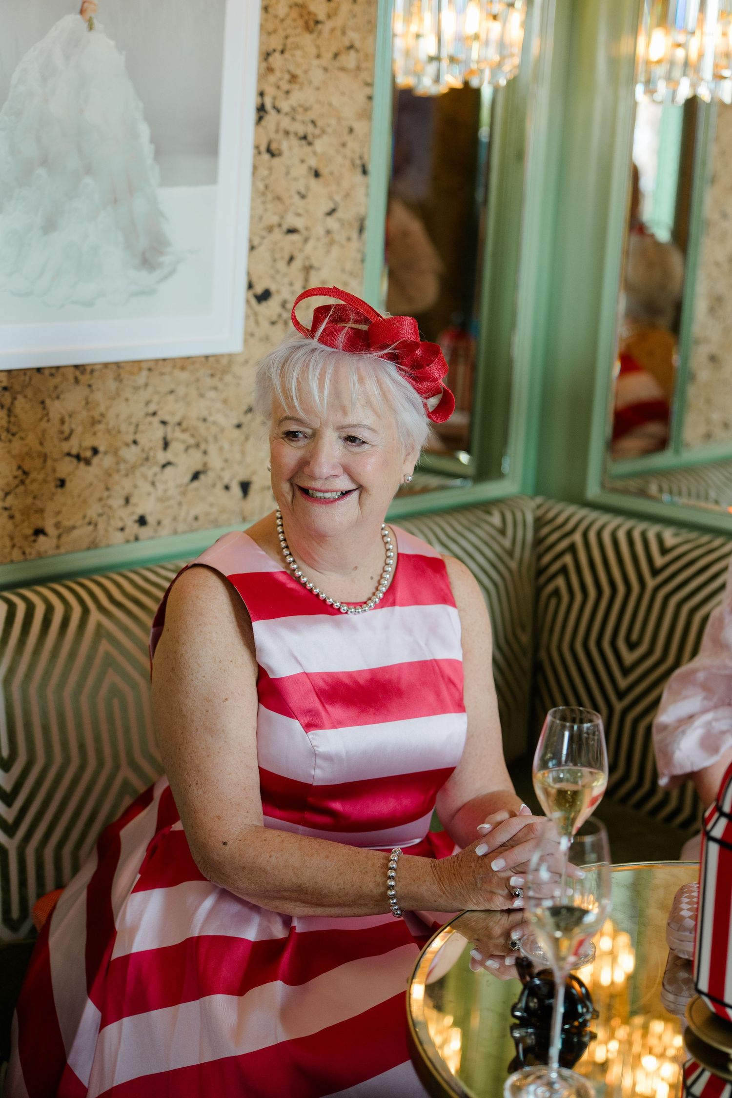 Elegant older woman in red and white striped dress with red fascinator holding champagne at wedding reception.