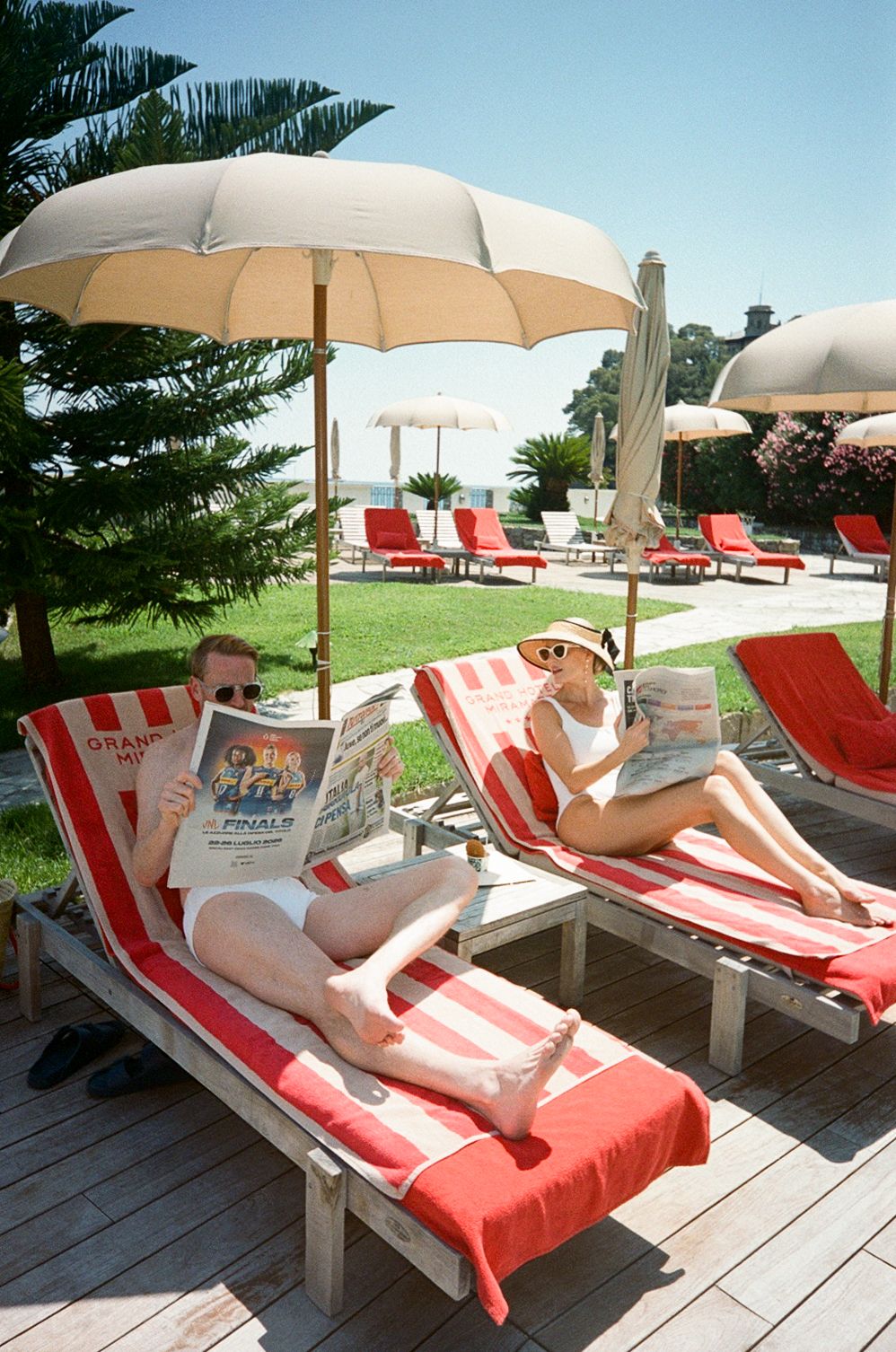 Couple relaxing on red striped sun loungers under beach umbrellas reading newspapers at a luxury Italian seaside resort.