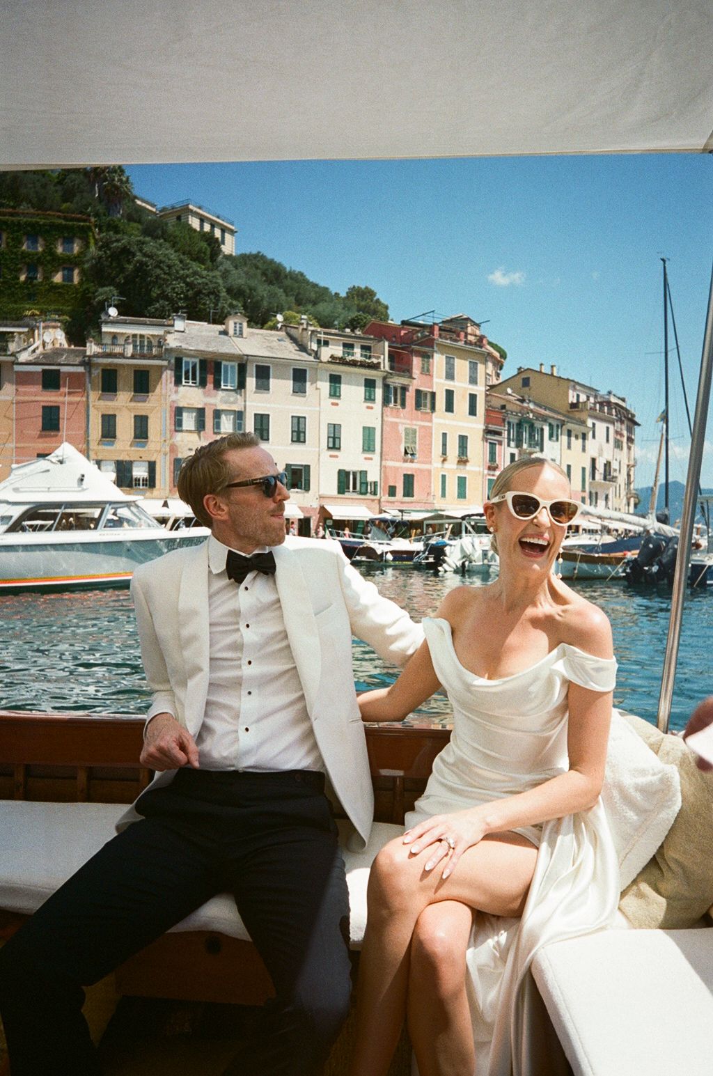Laughing bride in off-shoulder gown and groom in white tuxedo sitting on a boat with Portofino harbor behind them.