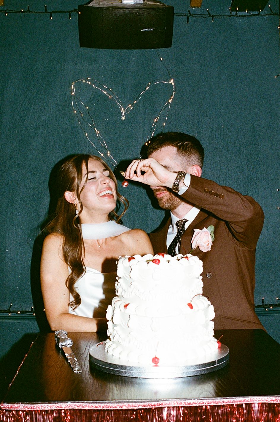 Laughing bride and groom cutting white wedding cake with heart light display in background.