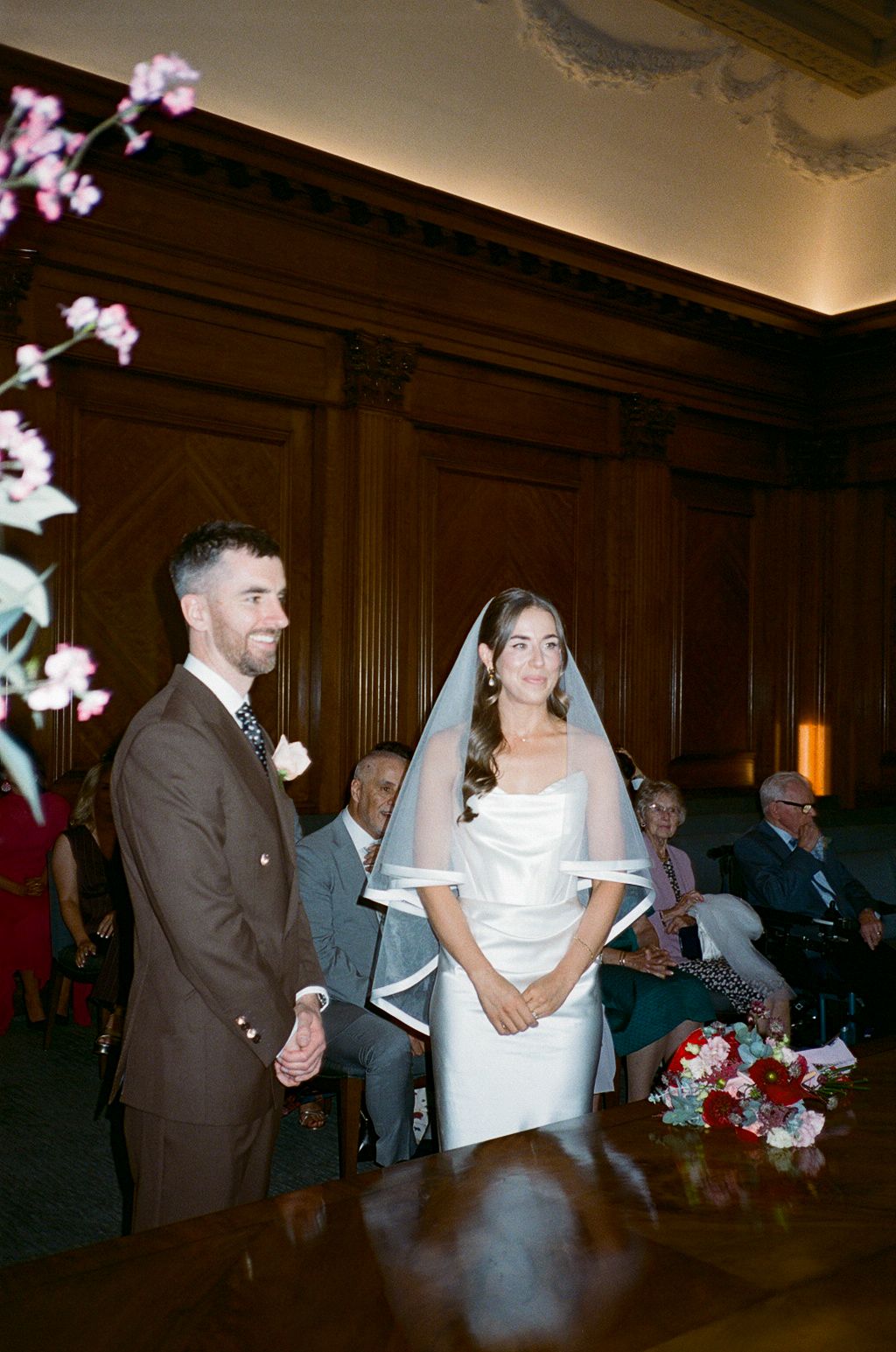 Bride in white gown and veil stands with groom in brown suit during wedding ceremony.