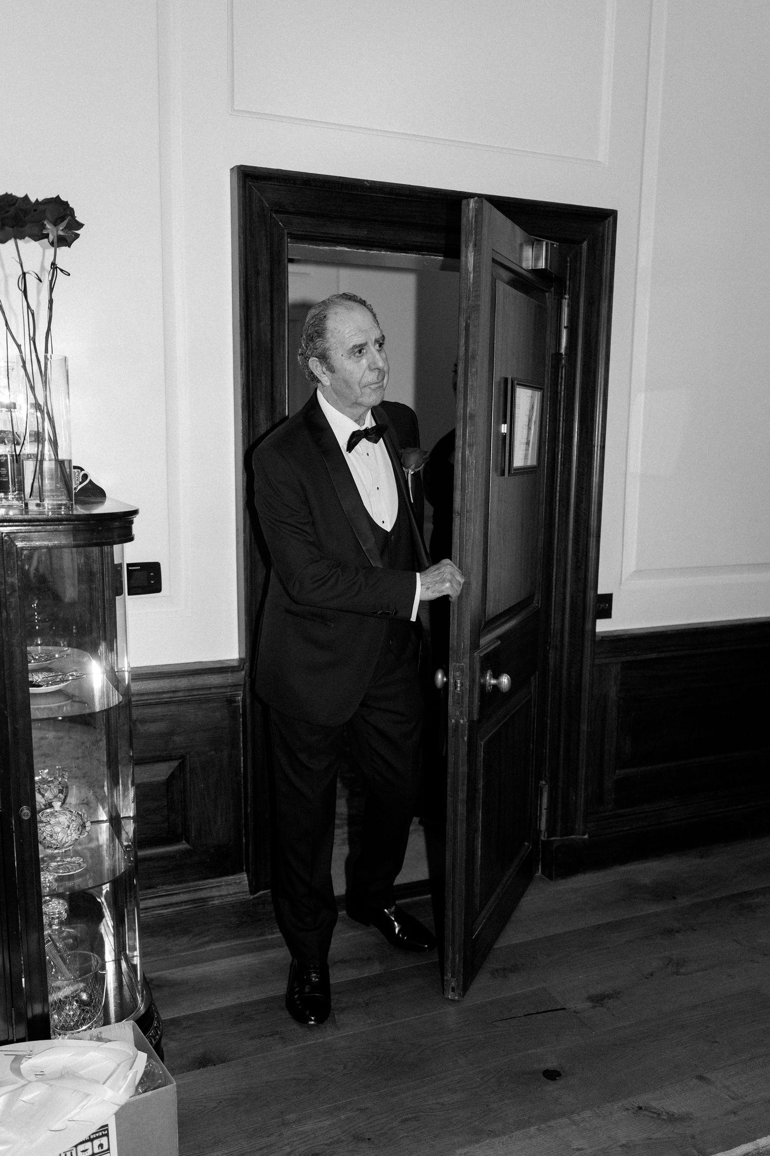 Black and white photo of a man in a tuxedo and bow tie stepping through an open wooden door into a paneled room.