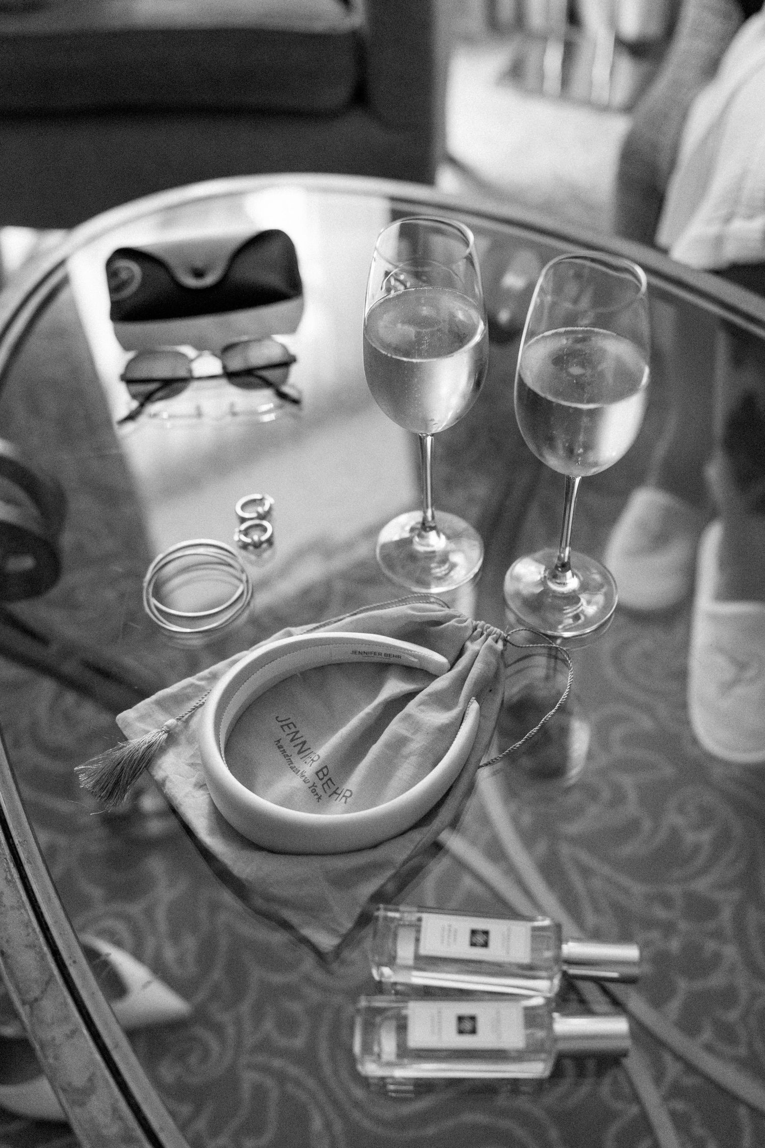 Wedding accessories including shoes, sunglasses, champagne glasses and perfume bottles on reflective surface.