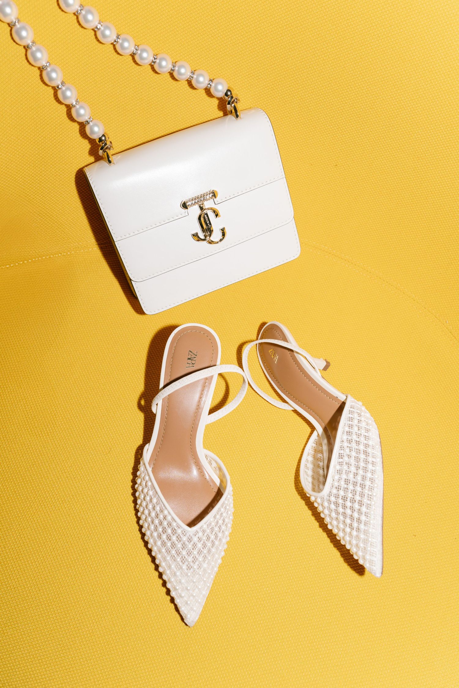 White designer shoes and handbag with pearl strap against yellow background.
