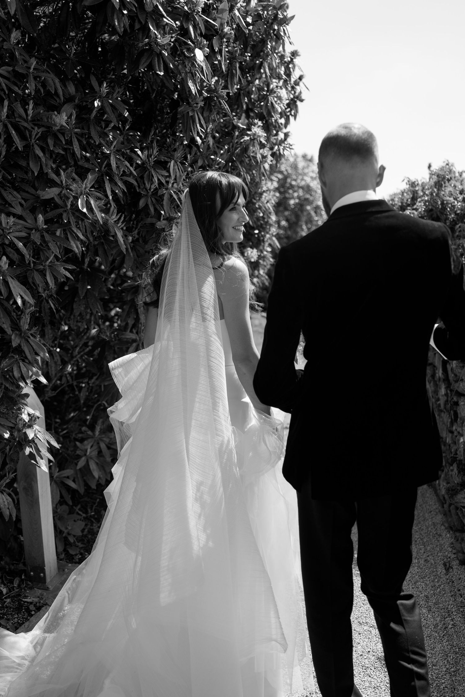 Elegant wedding procession walking through a garden path in black and white.