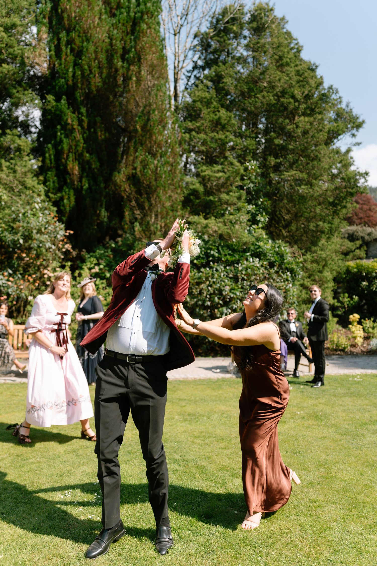 Wedding guests perform dynamic dance moves on a sunny lawn with trees in the background.