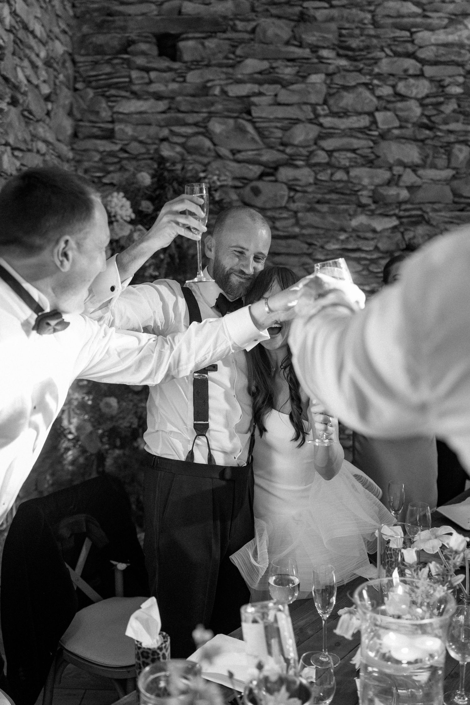 Wedding party guests raising glasses in a celebratory toast against a stone wall.