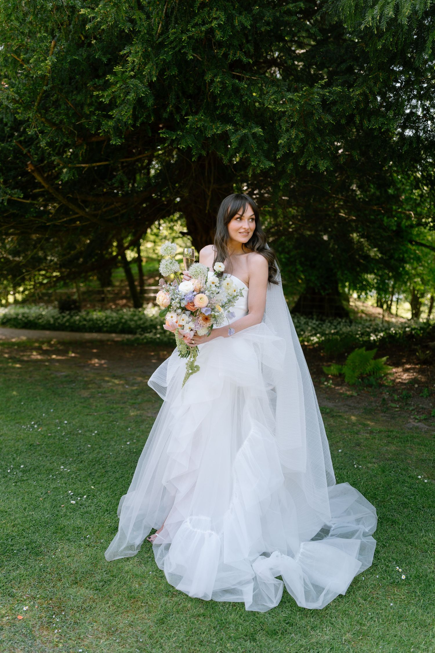 Bride in flowing white gown poses with bouquet on manicured lawn.