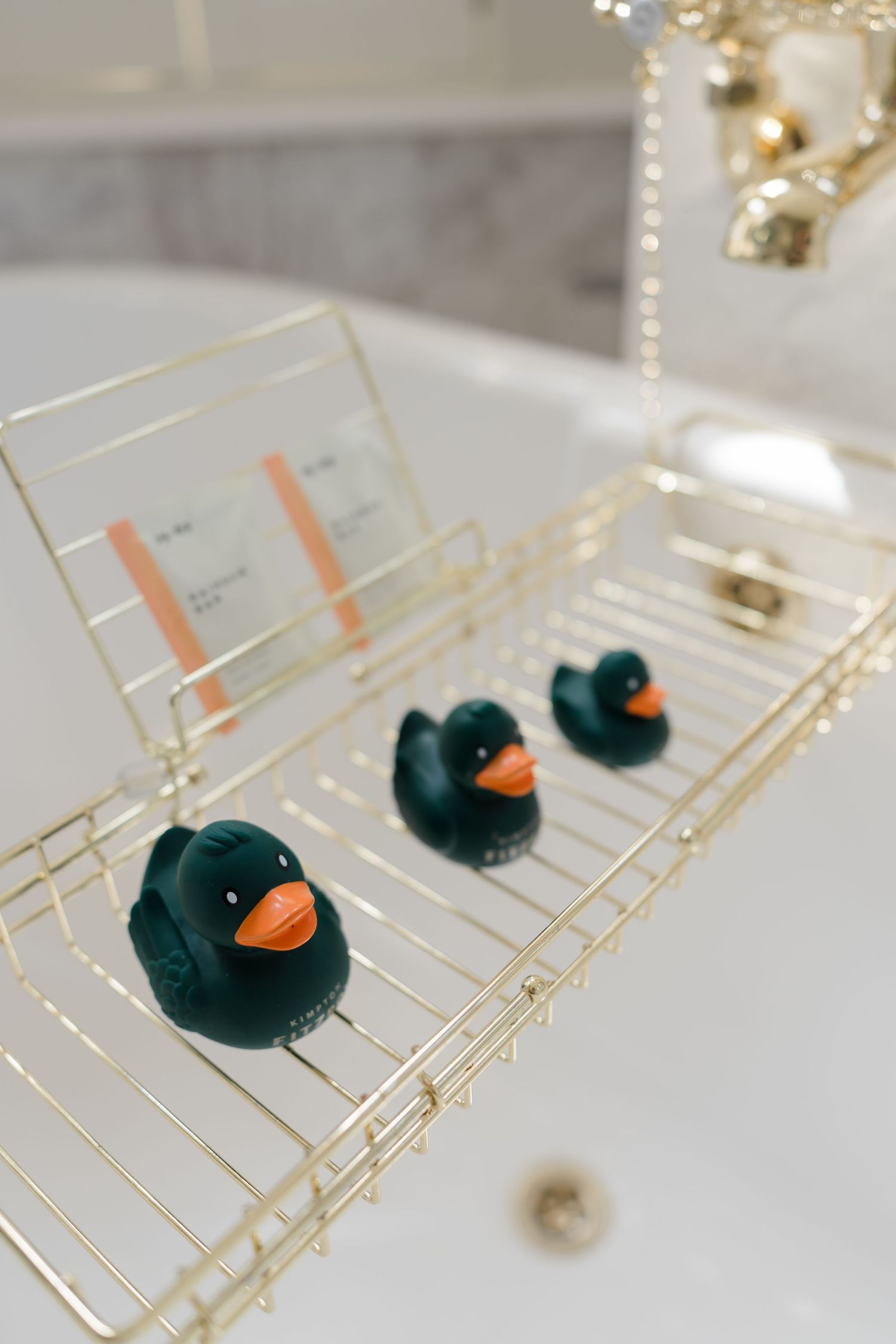 Three dark green rubber ducks on a gold wire bath caddy tray over a white bathtub with gold faucet.
