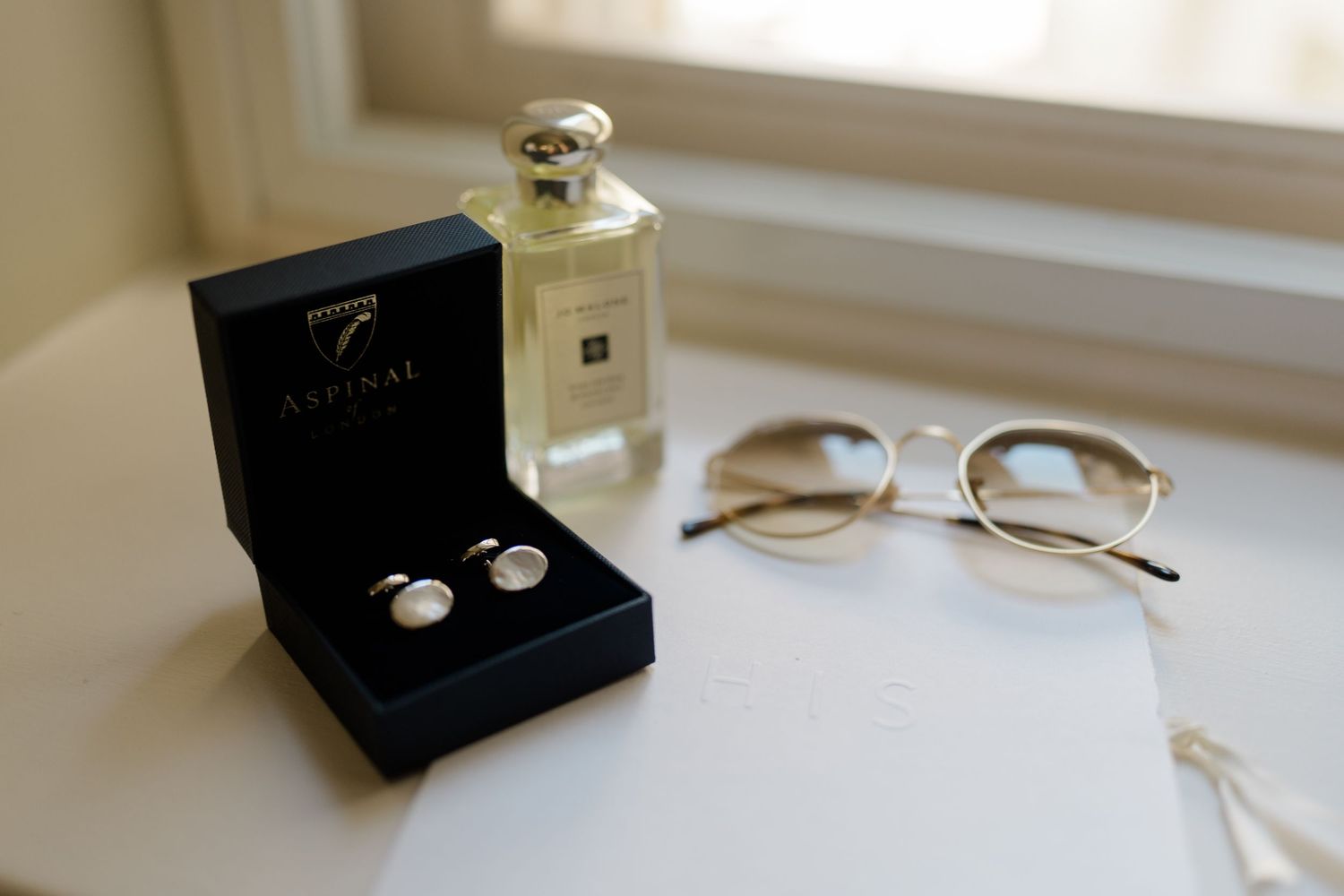 Aspinal cufflinks in black gift box beside Jo Malone perfume and gold-rimmed glasses on white surface.