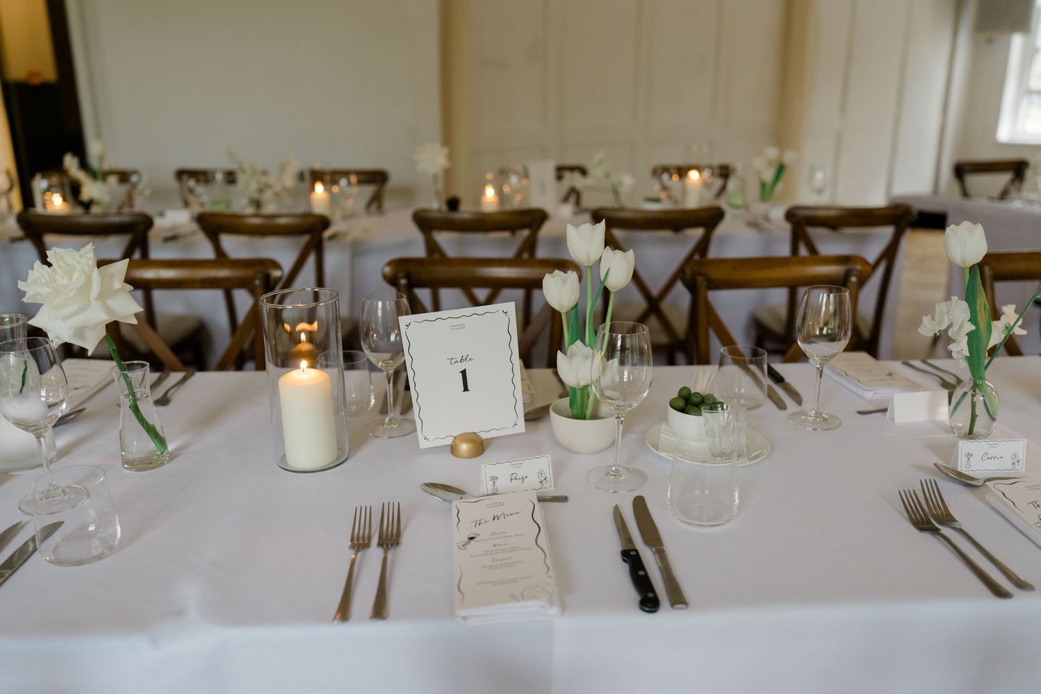 Elegant wedding reception table set with white tulips, candles, and gold cutlery in a bright venue.