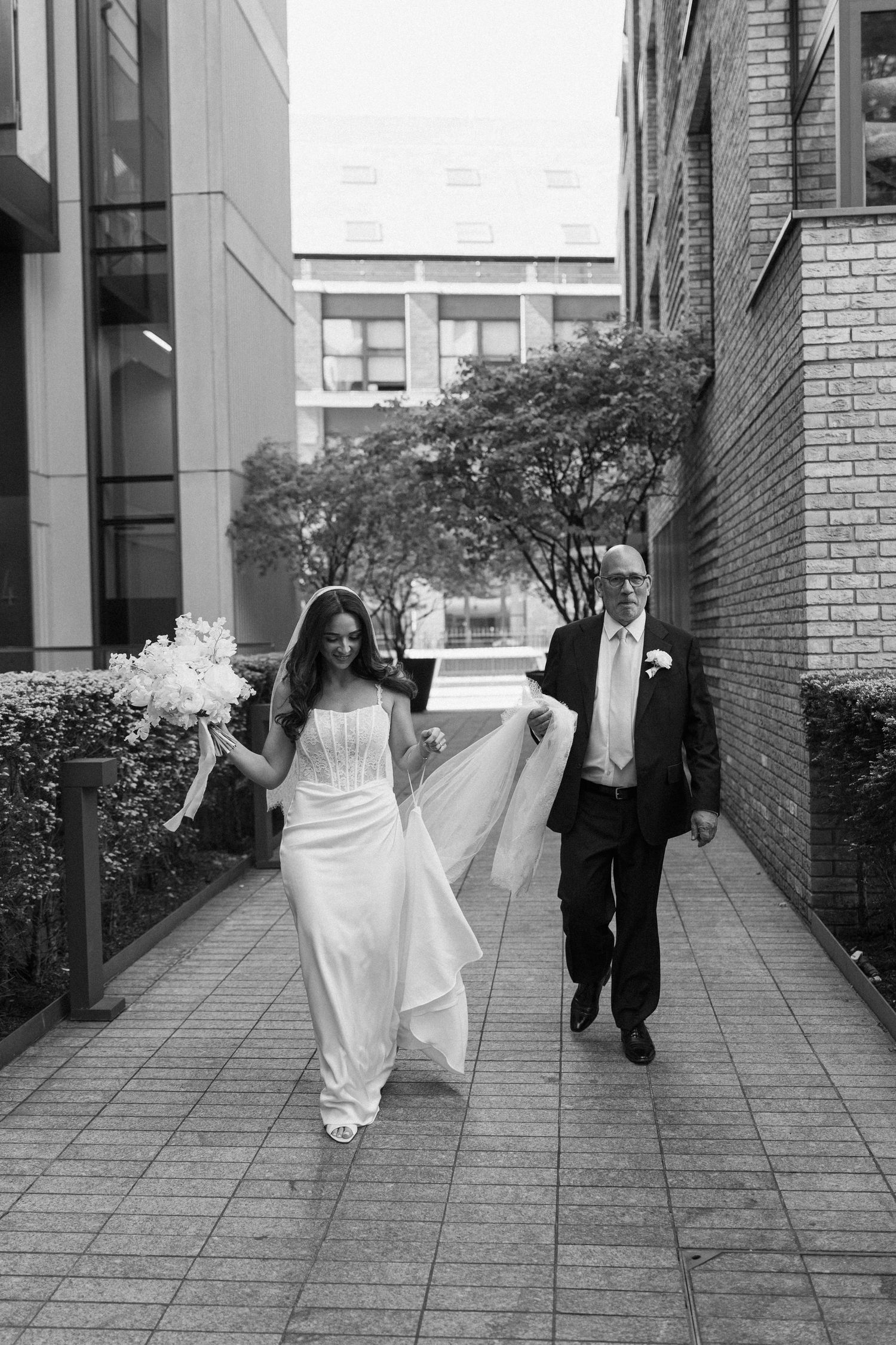 Black and white photo of bride in white gown walking with her father along a city pathway.