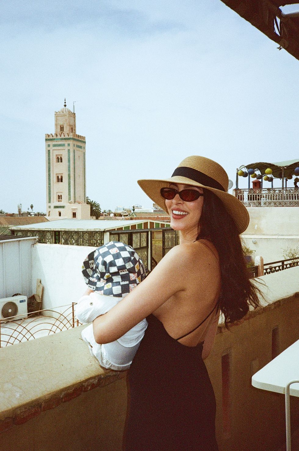 Woman in straw hat and sunglasses holding baby on rooftop terrace with mosque minaret in background.