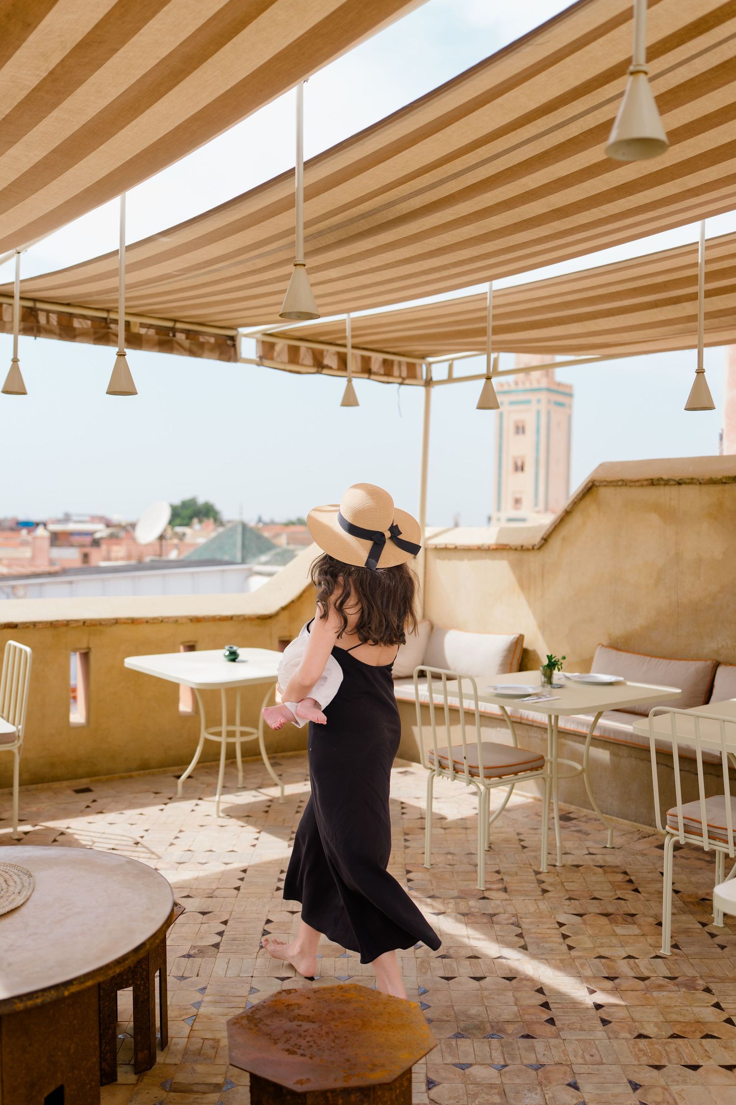 Woman in black dress and straw hat on a Moroccan rooftop terrace with striped awning and city views.