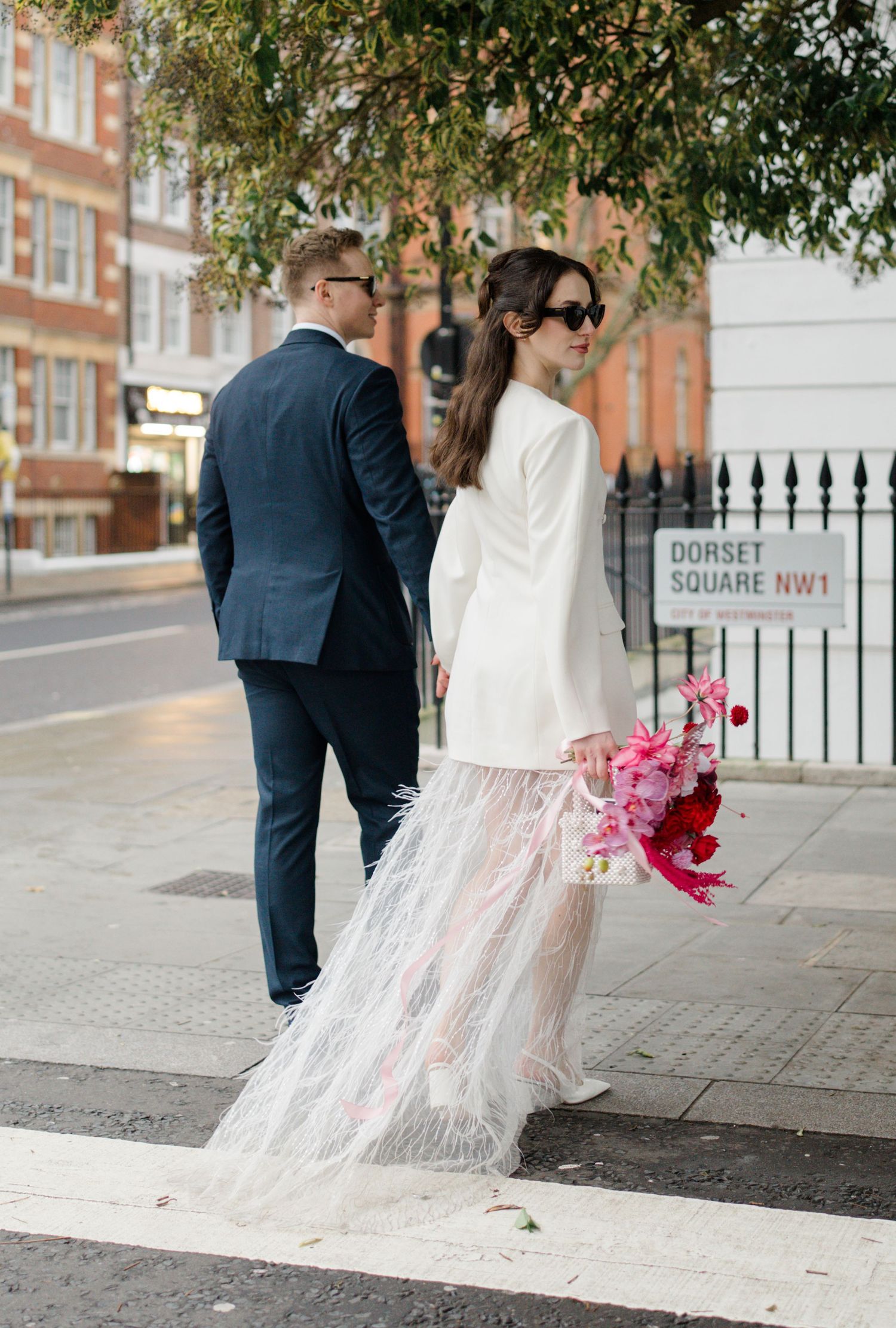 Bride in white blazer and feathered skirt with groom crossing street at Dorset Square London.