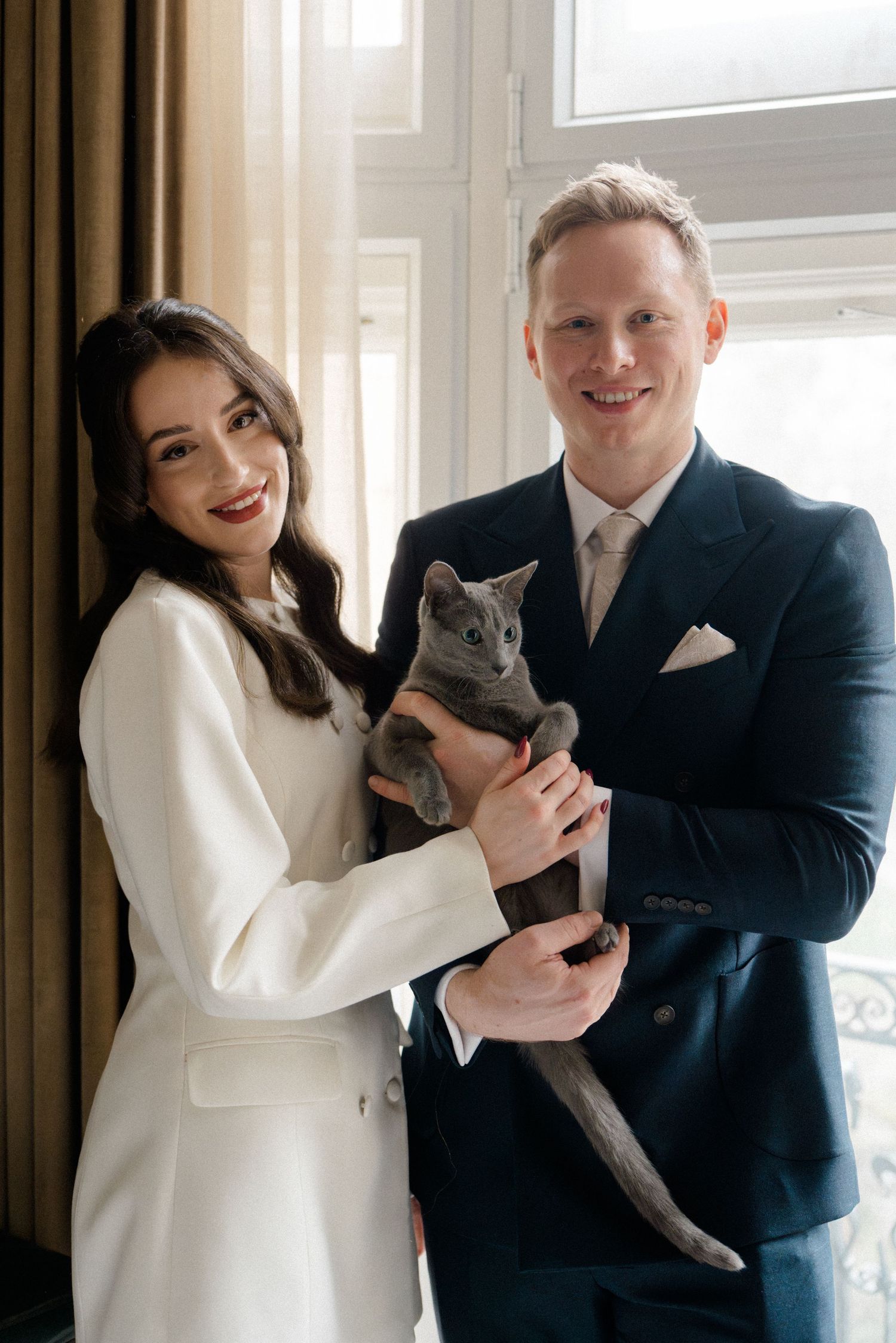 Smiling bride and groom in white suit and navy tuxedo hold a gray Russian Blue cat.