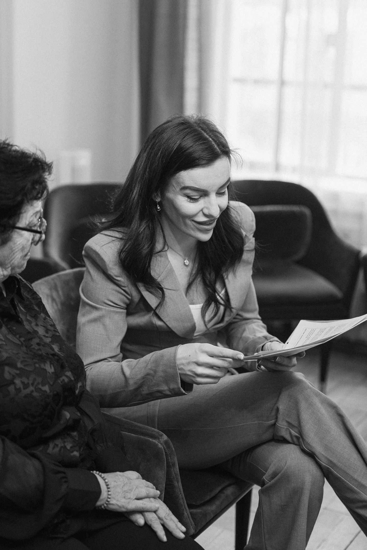 Black and white photo of smiling dark-haired woman in suit reviewing documents with elderly woman while seated indoors.