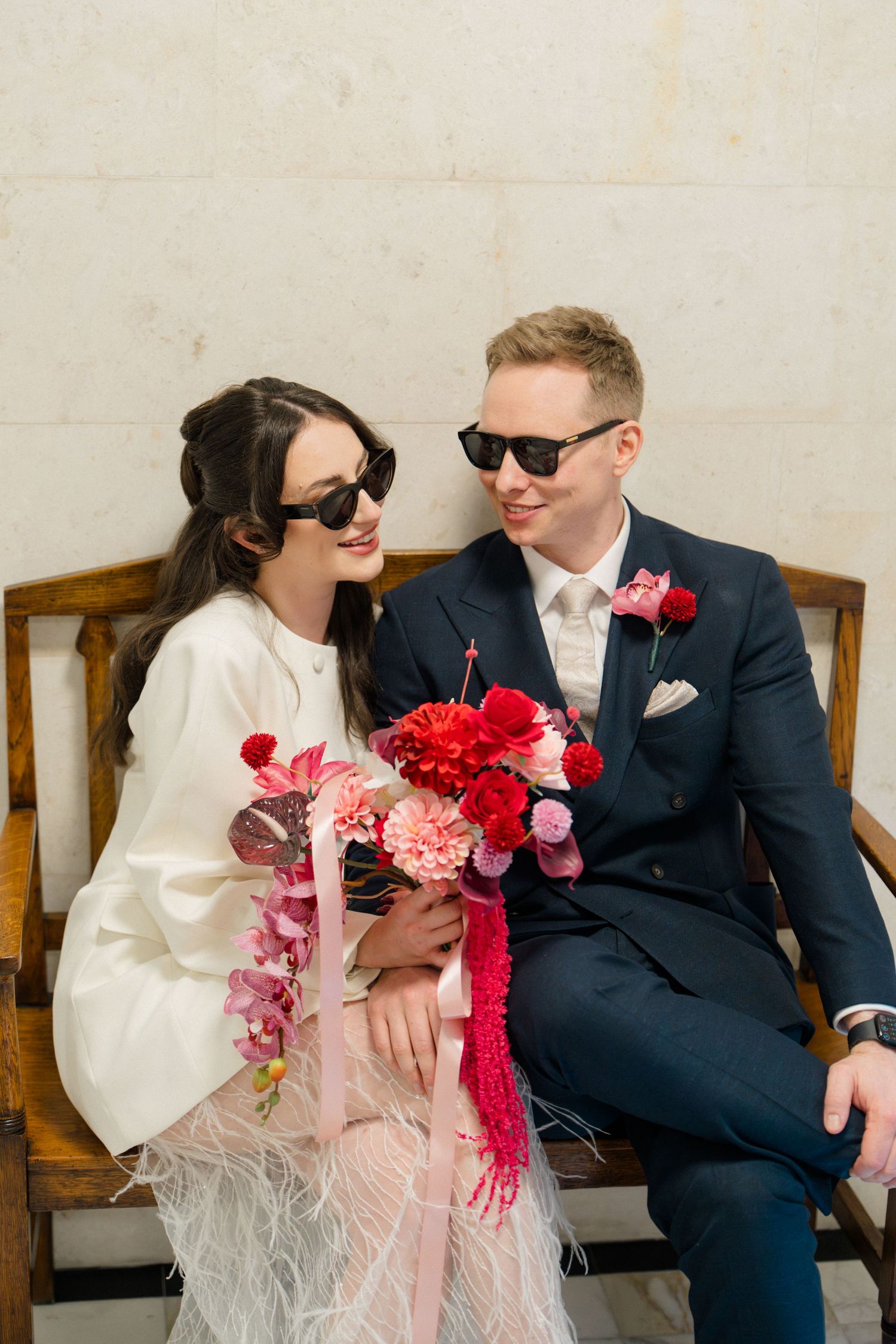 Bride and groom in sunglasses sit together holding a vibrant pink and red bouquet.