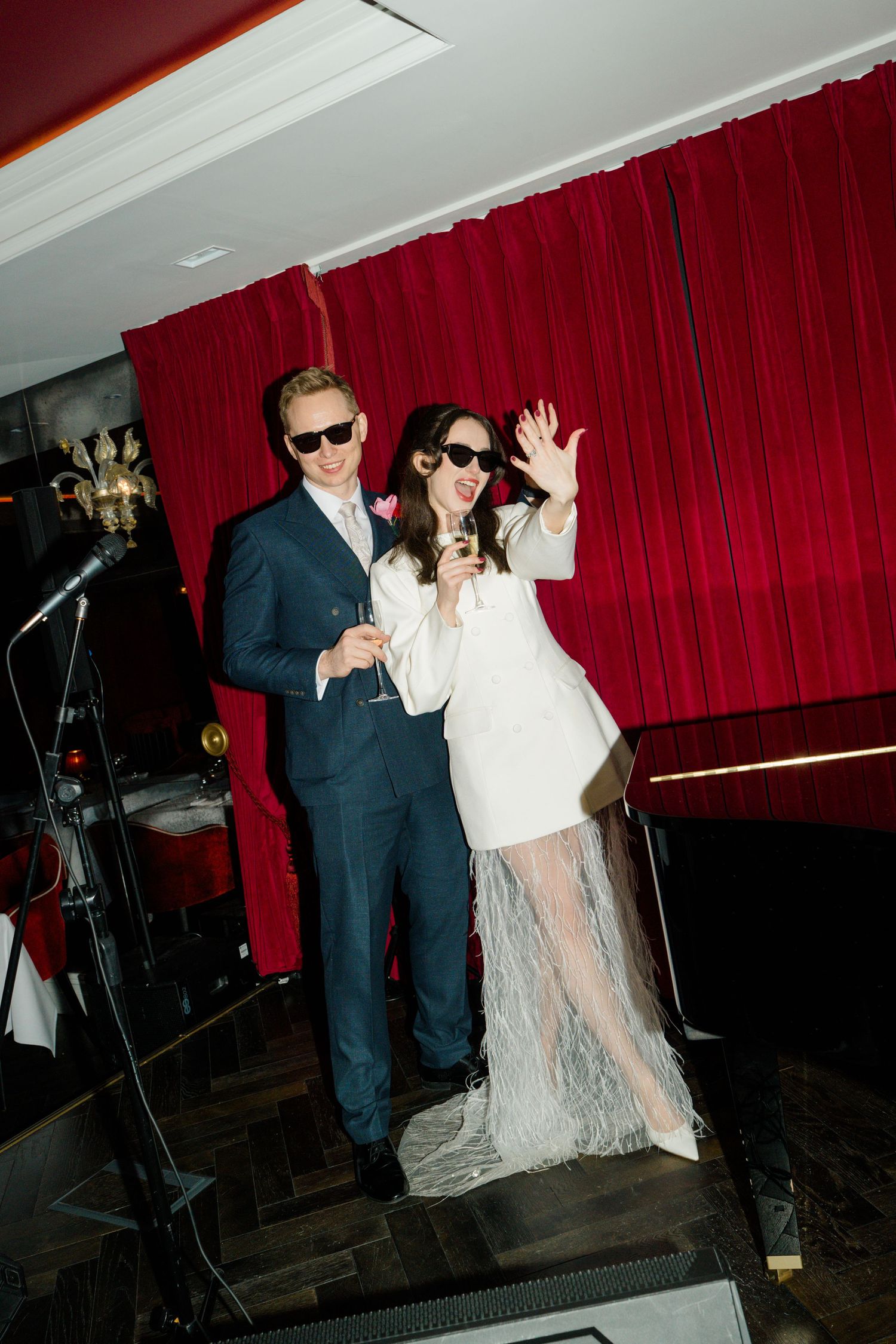 Bride and groom wearing sunglasses celebrating at wedding reception with red curtain backdrop.