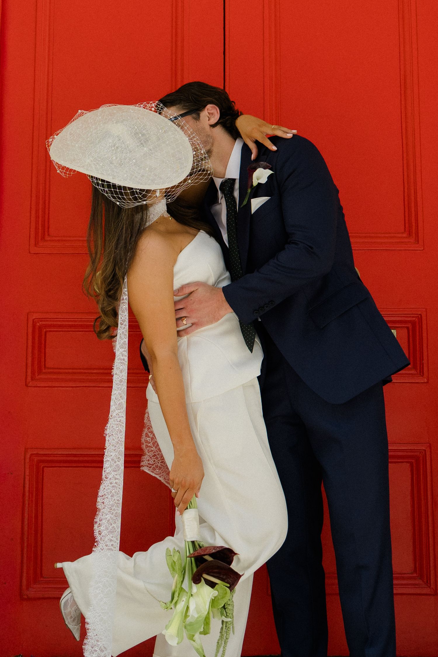 Bride in white gown and fascinator kisses groom in navy suit against a vibrant red door, holding a green bouquet.