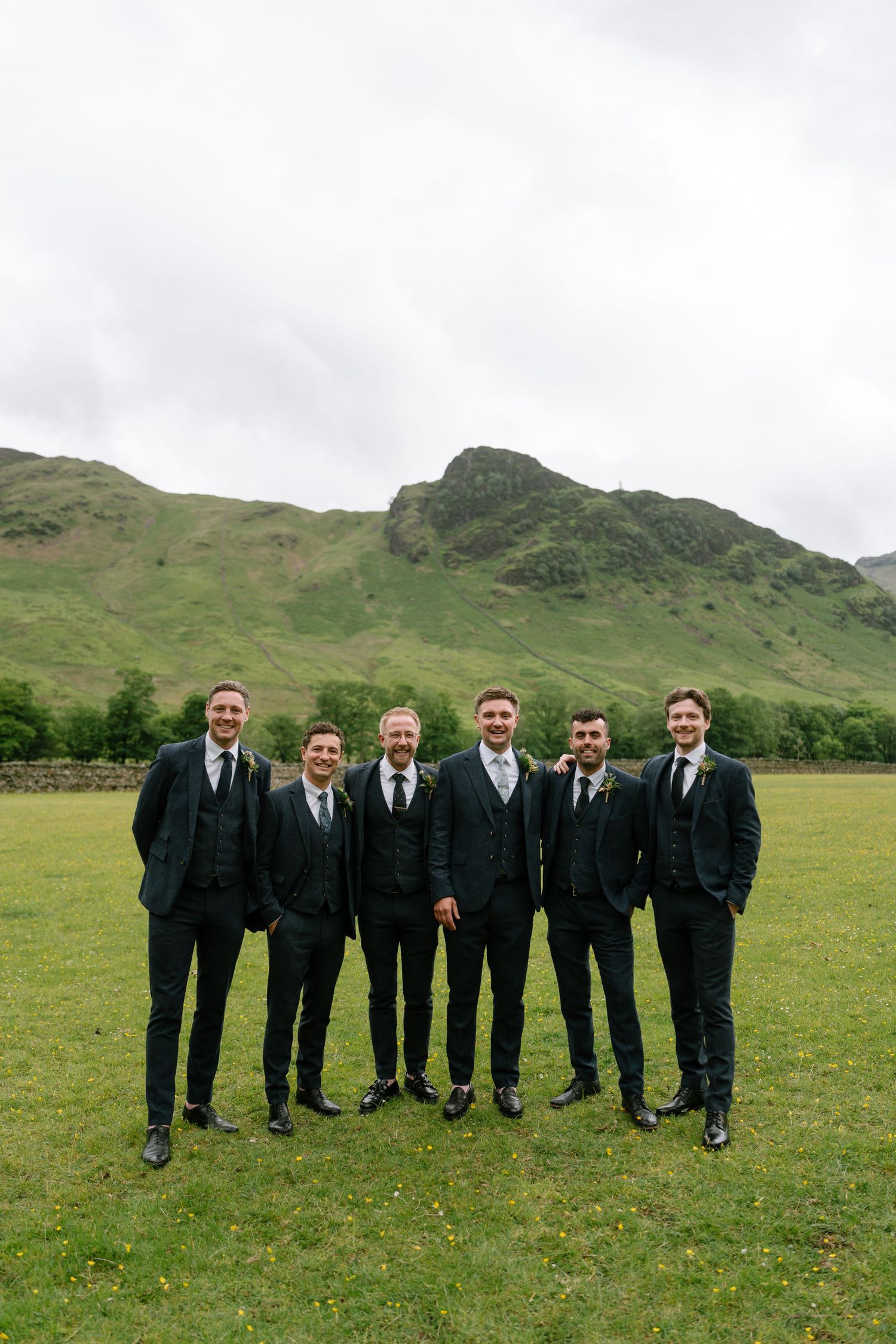 Groomsmen in dark suits standing on grass with rolling hills in the background.