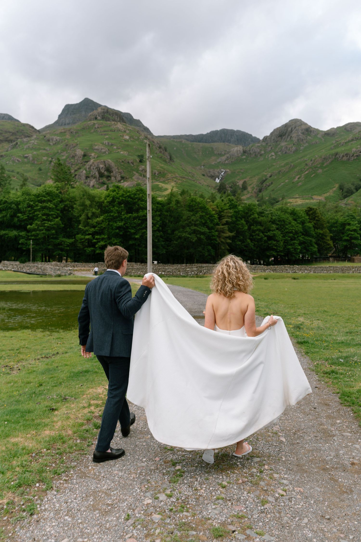 A couple walks along a mountain path with dramatic peaks and green hills in the background on their wedding day.
