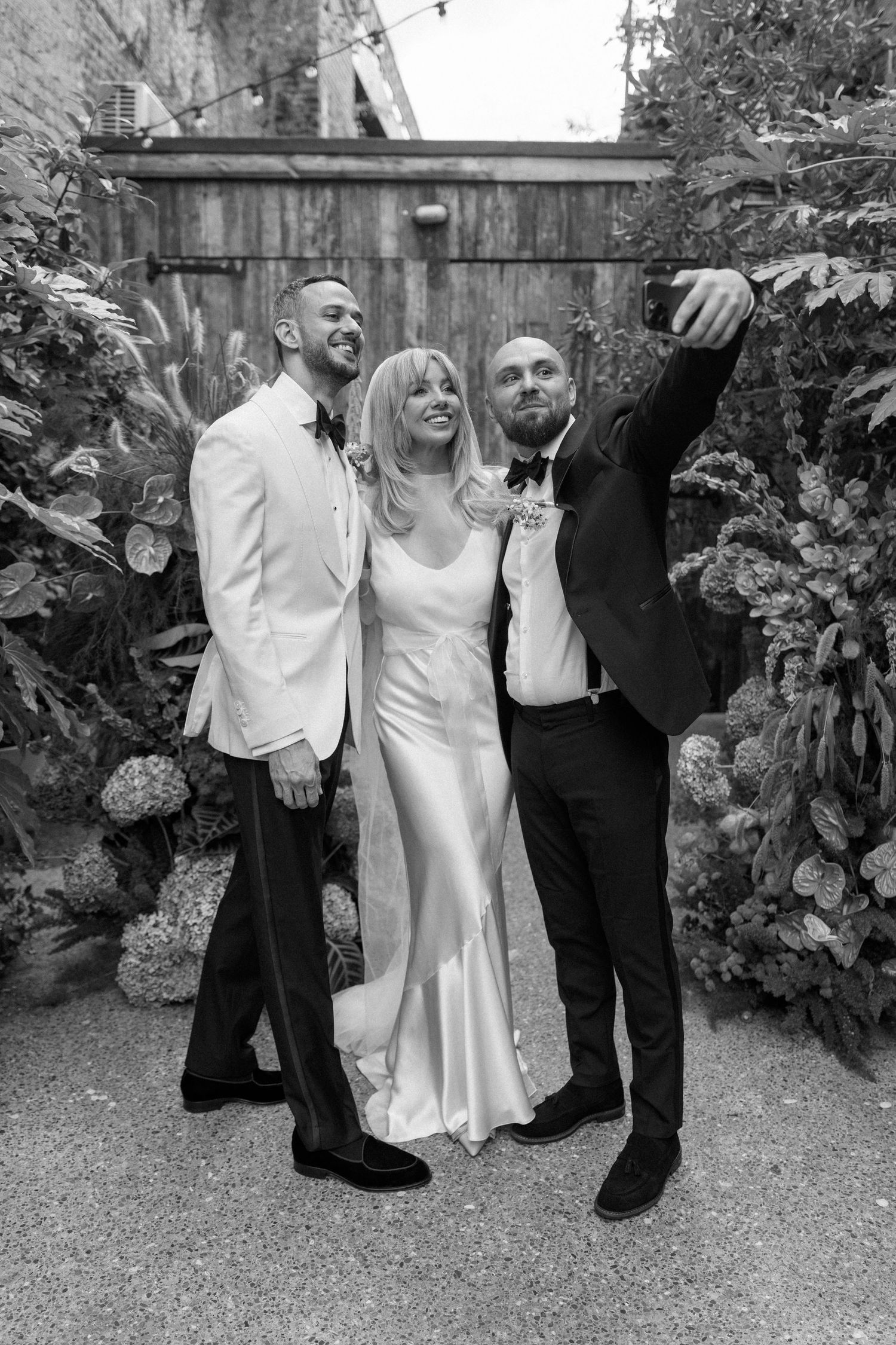 Wedding party in black and white attire posing for photos in garden setting.