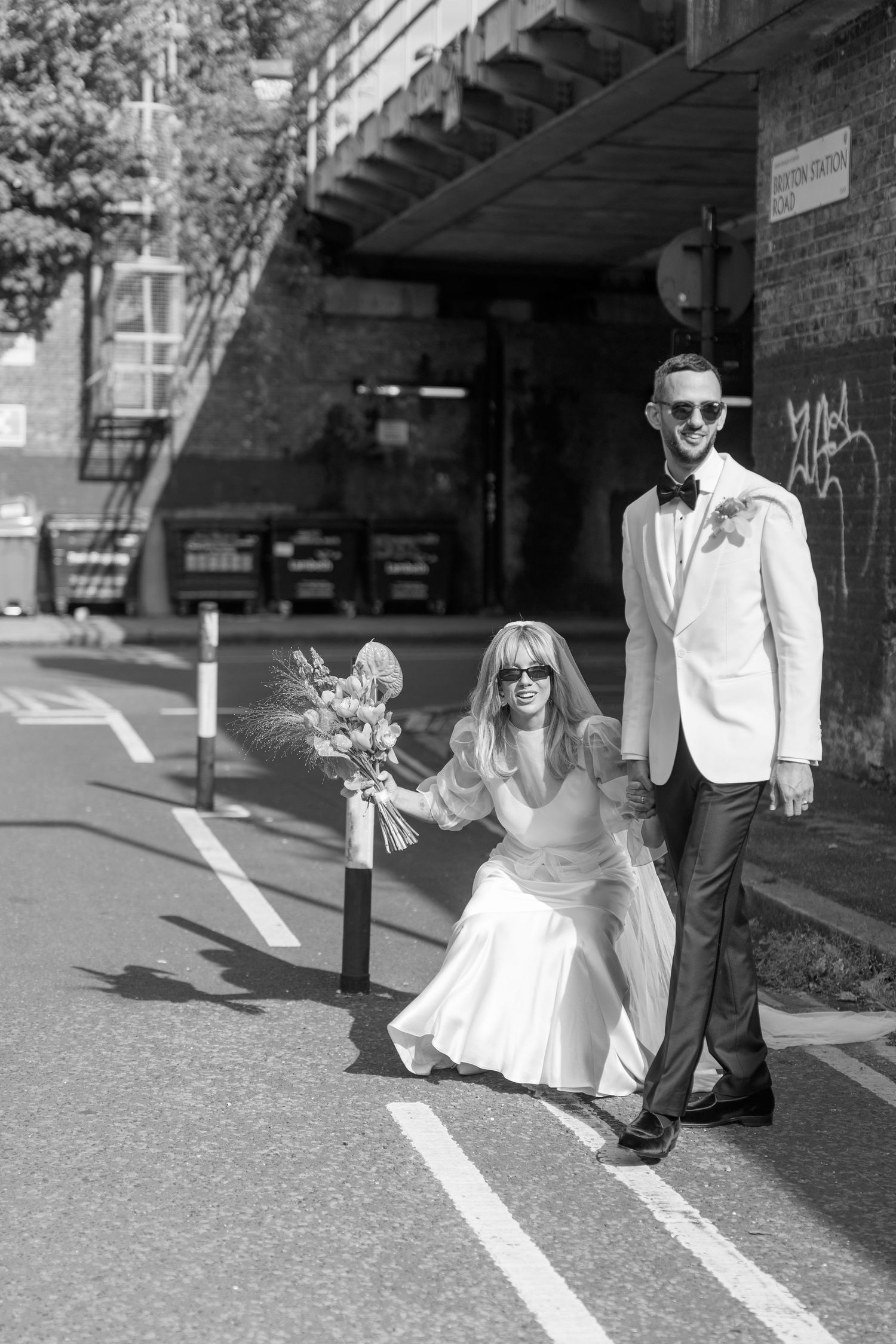 Couple in wedding attire poses playfully on painted street lines in black and white.
