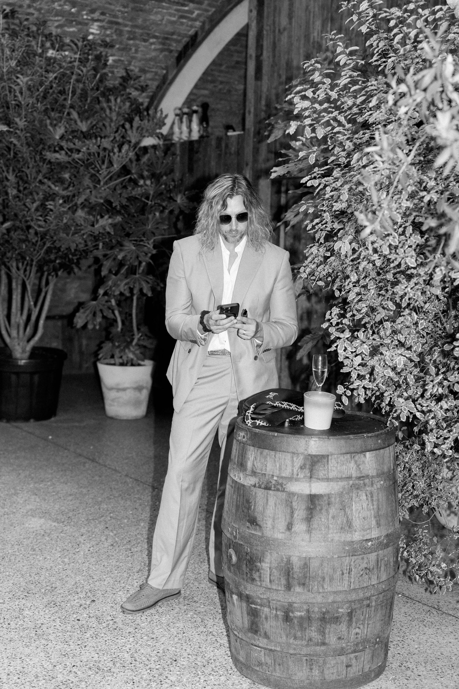 Person in sunglasses and light suit poses near wooden barrel in garden setting.