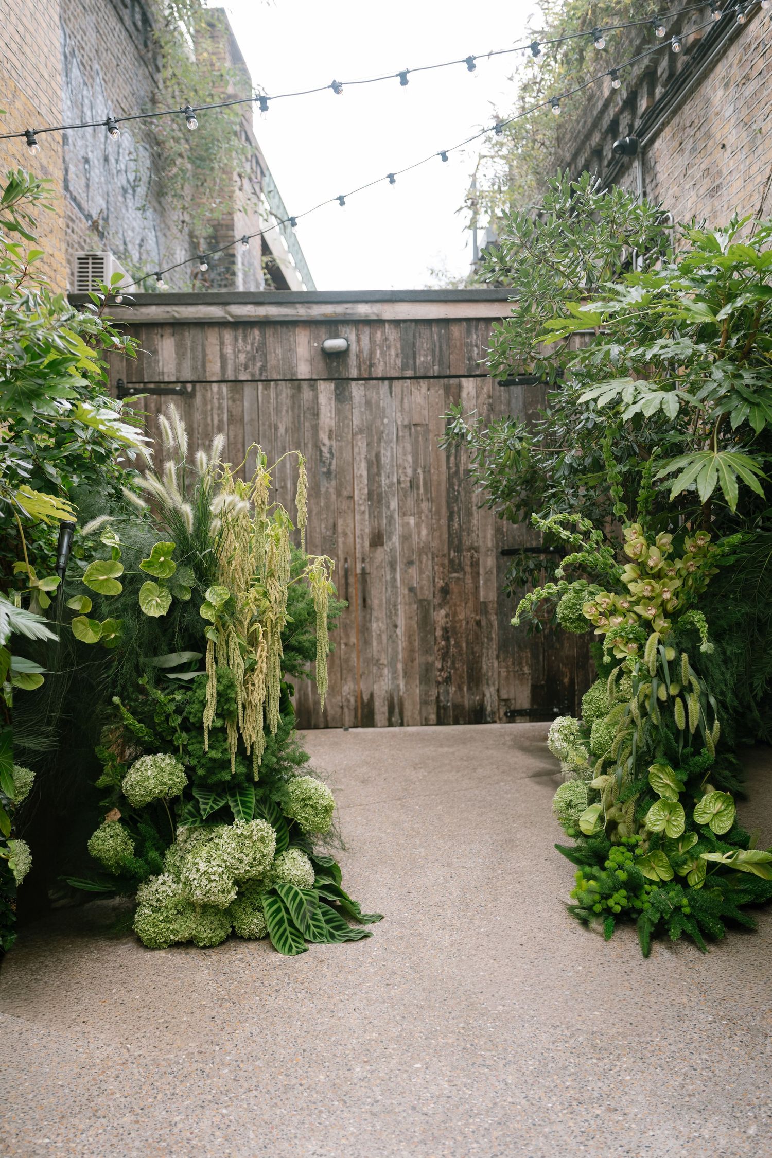 Outdoor garden venue with string lights and wooden fence surrounded by lush greenery.