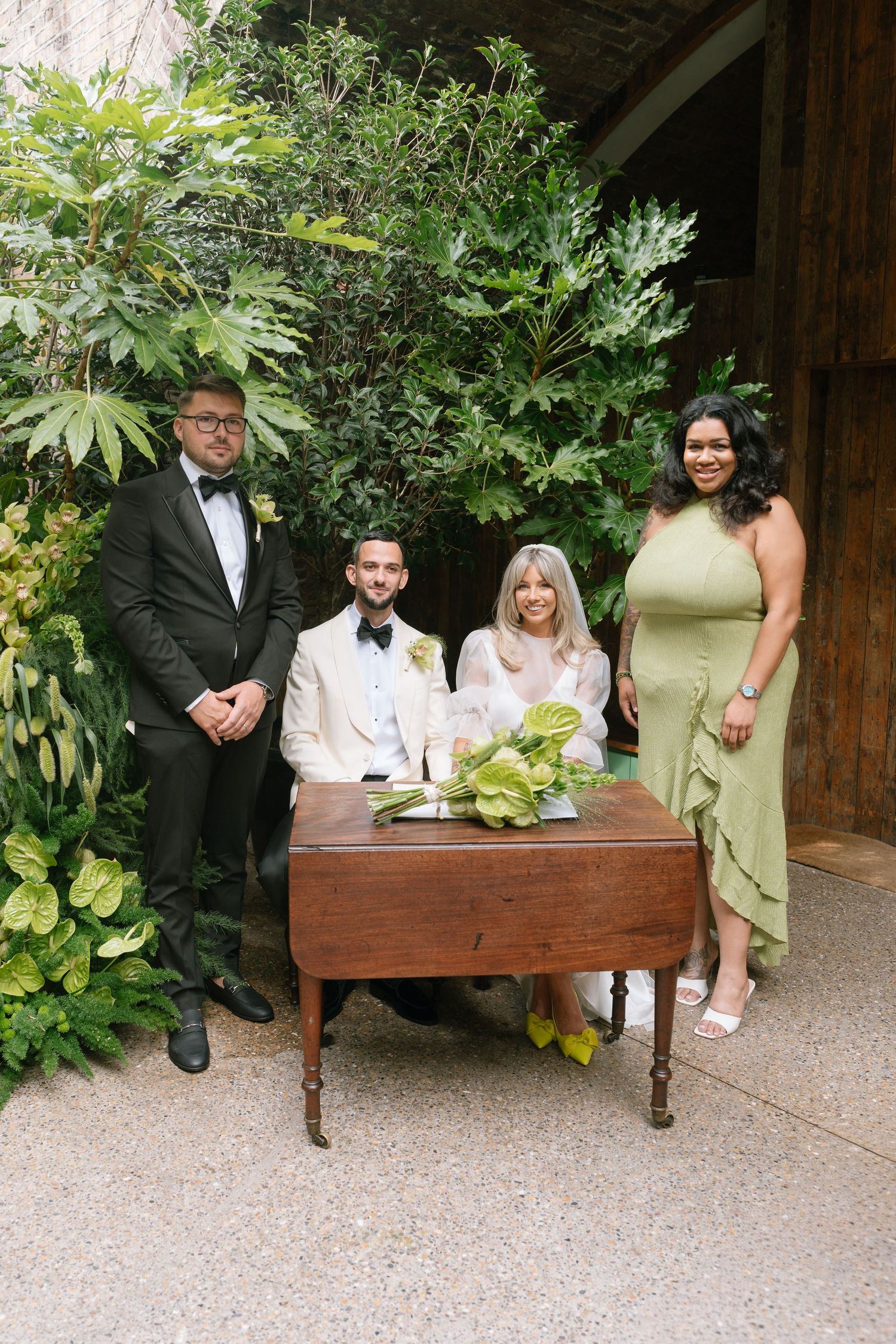 Wedding party members pose casually on a wooden bench in a garden setting.