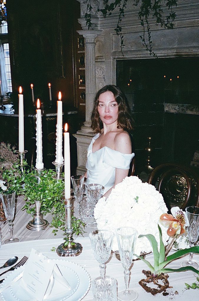 Elegant dinner table setting with white candles, greenery, and vintage decor in moody lighting.