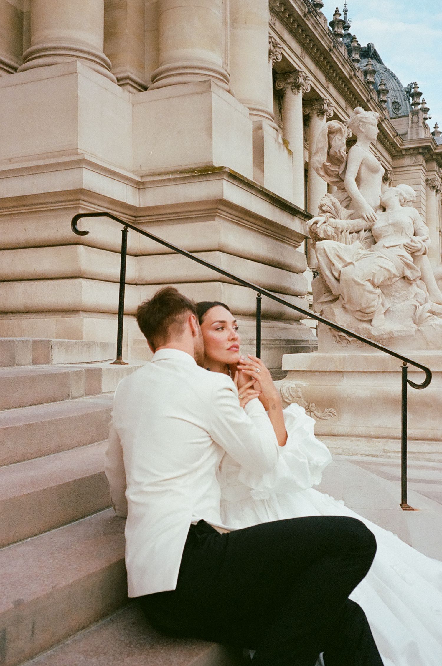 A romantic couple in wedding attire embraces on elegant stone steps of a grand building with classical architecture.