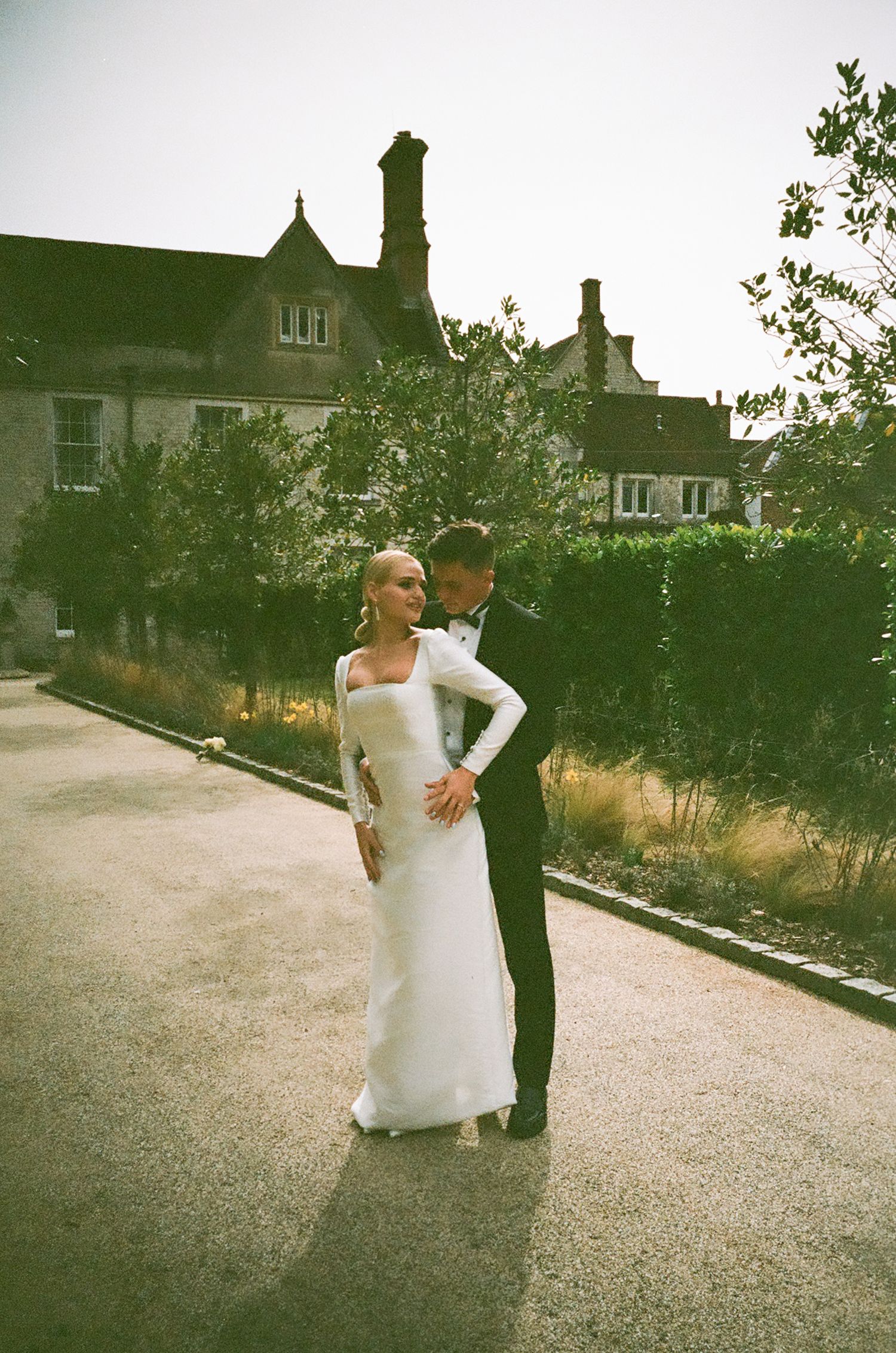 A couple in wedding attire embrace on a path with a traditional English house in the background.