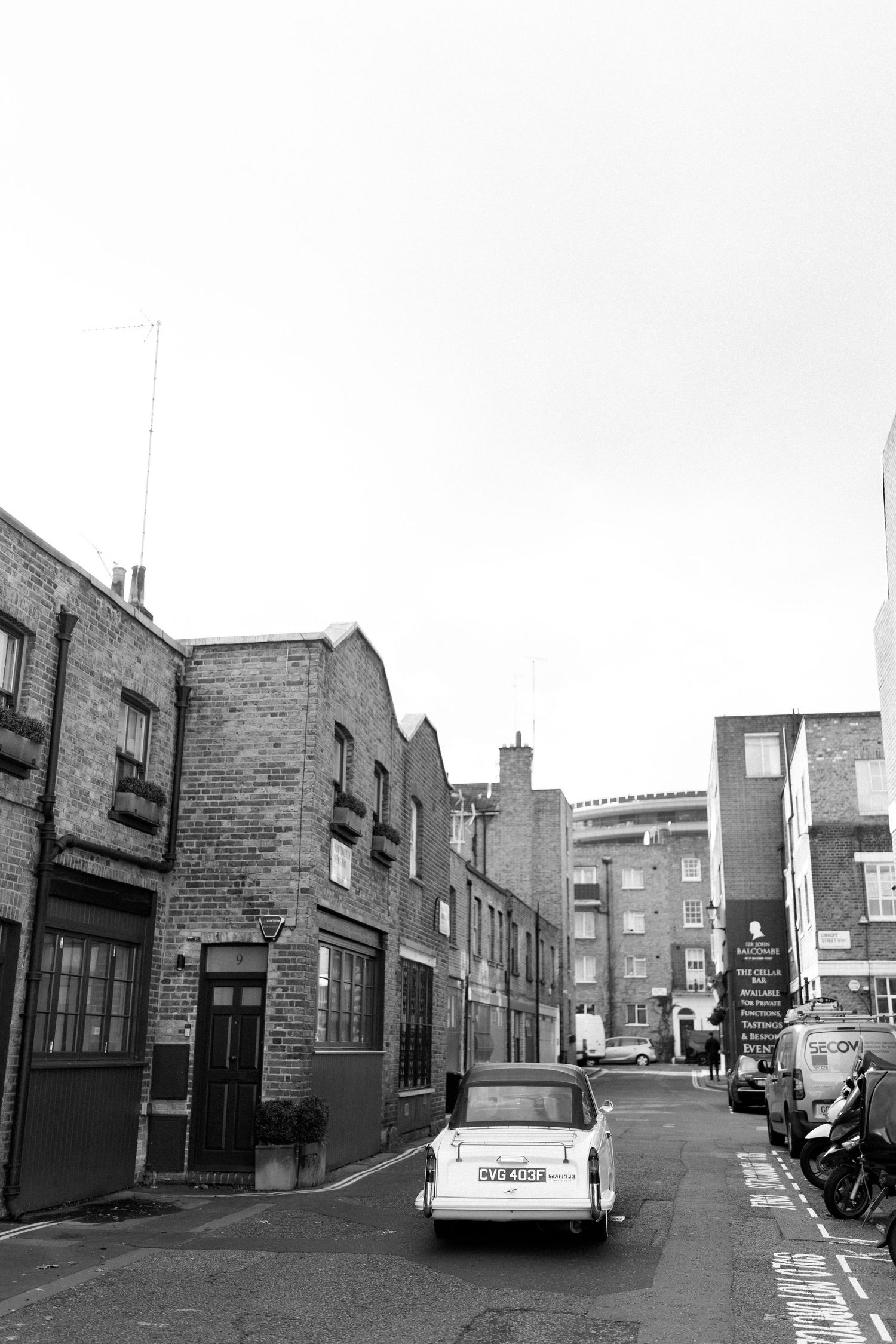 Vintage black and white street scene showing brick buildings and a classic car parked on a narrow urban road.