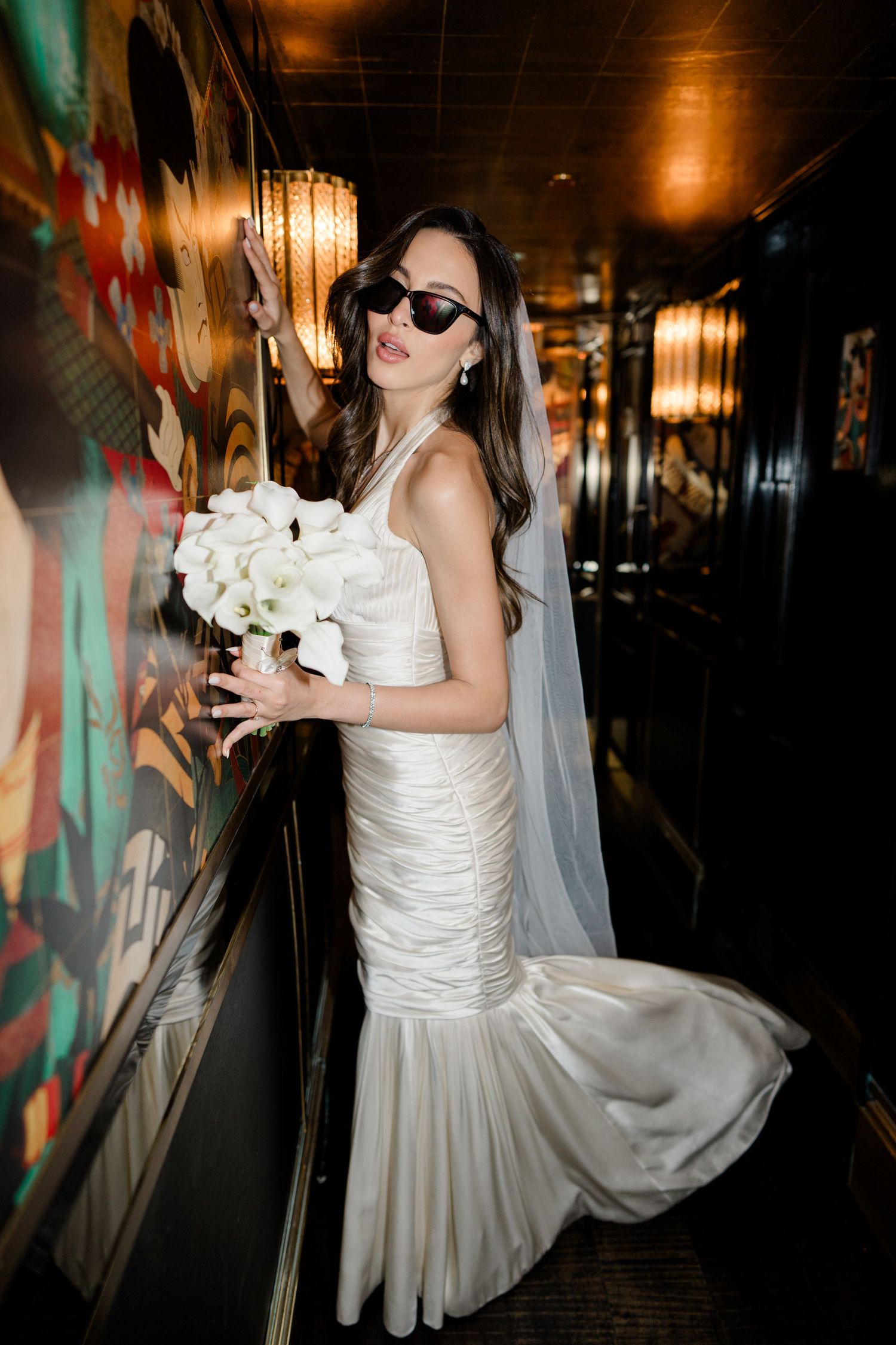 A bride in a white dress holds a bouquet in a dimly lit hallway with artistic wall murals.