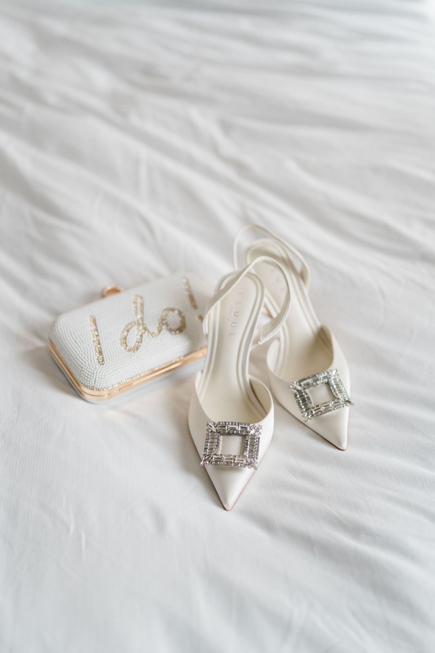 Elegant white bridal shoes with crystal buckles displayed on white bedding.