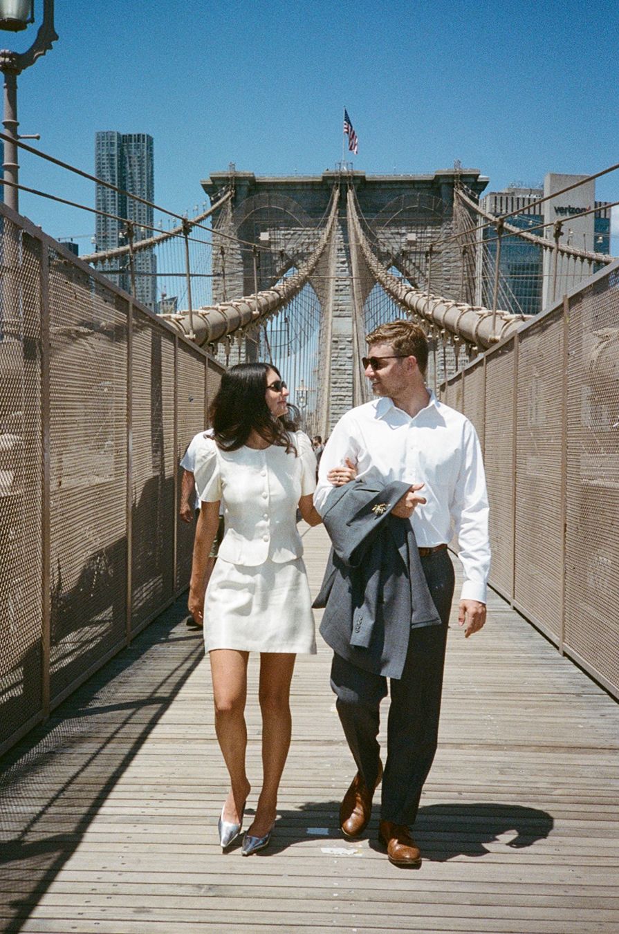 Couple walking hand-in-hand on Brooklyn Bridge pedestrian walkway on a sunny day, Manhattan skyline visible behind them.