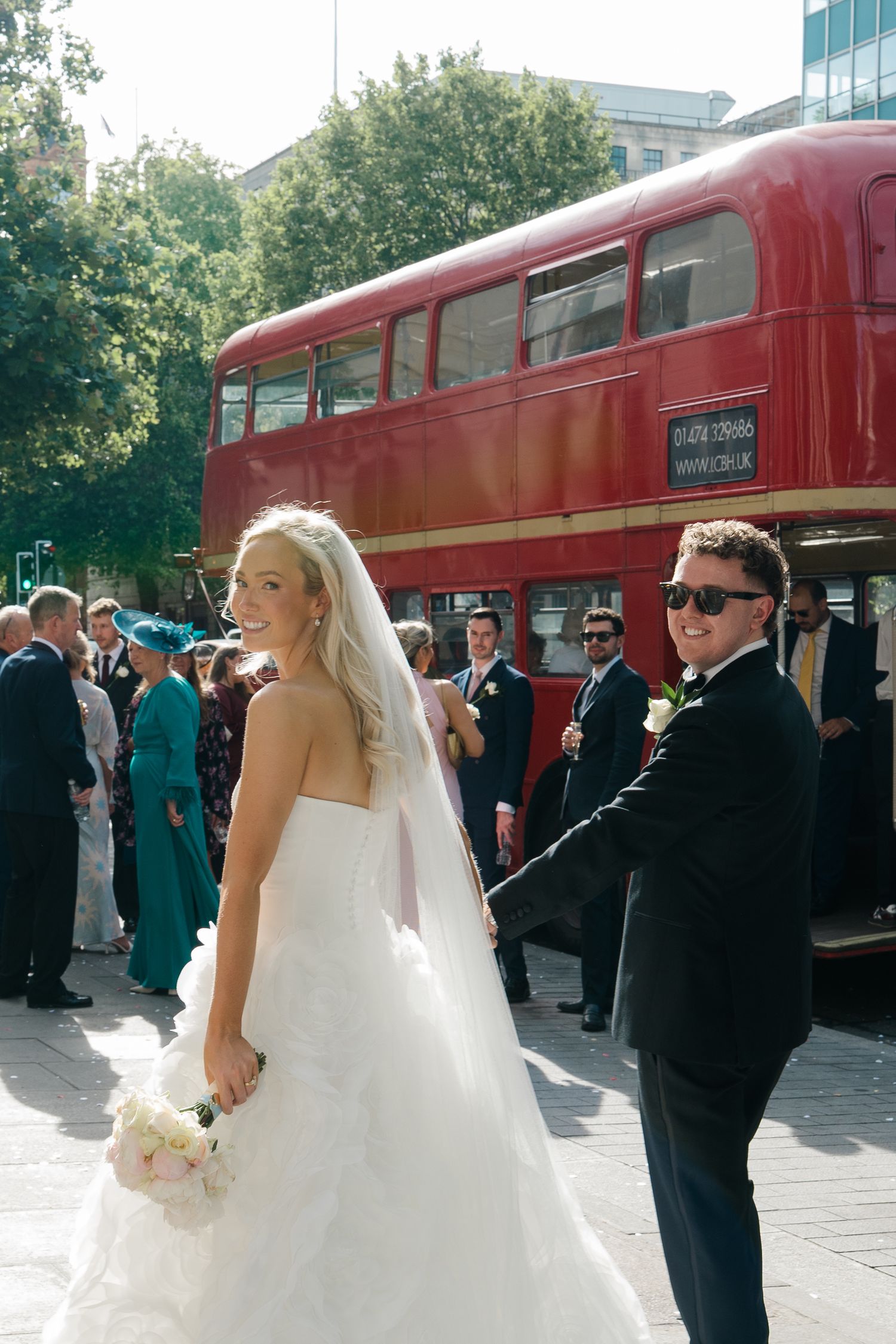 Bride in white gown and groom in black suit pose near a red double-decker bus with wedding guests in London.