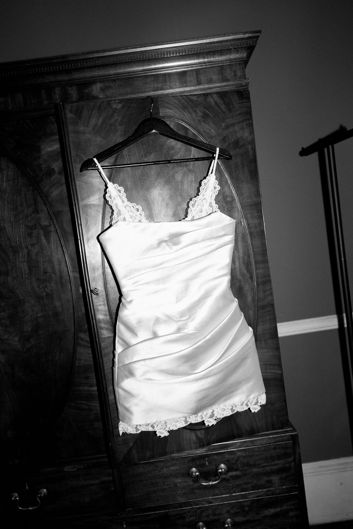 White lace spaghetti strap wedding dress hanging on wooden armoire in black and white photo.