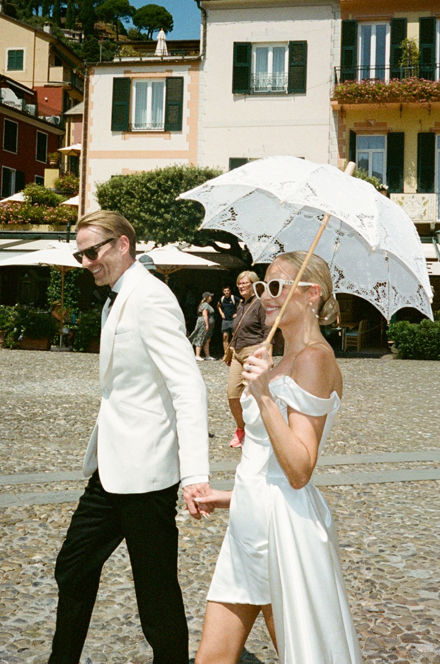 Bride and groom walking through a sunlit Italian piazza, bride holding a white lace parasol umbrella.