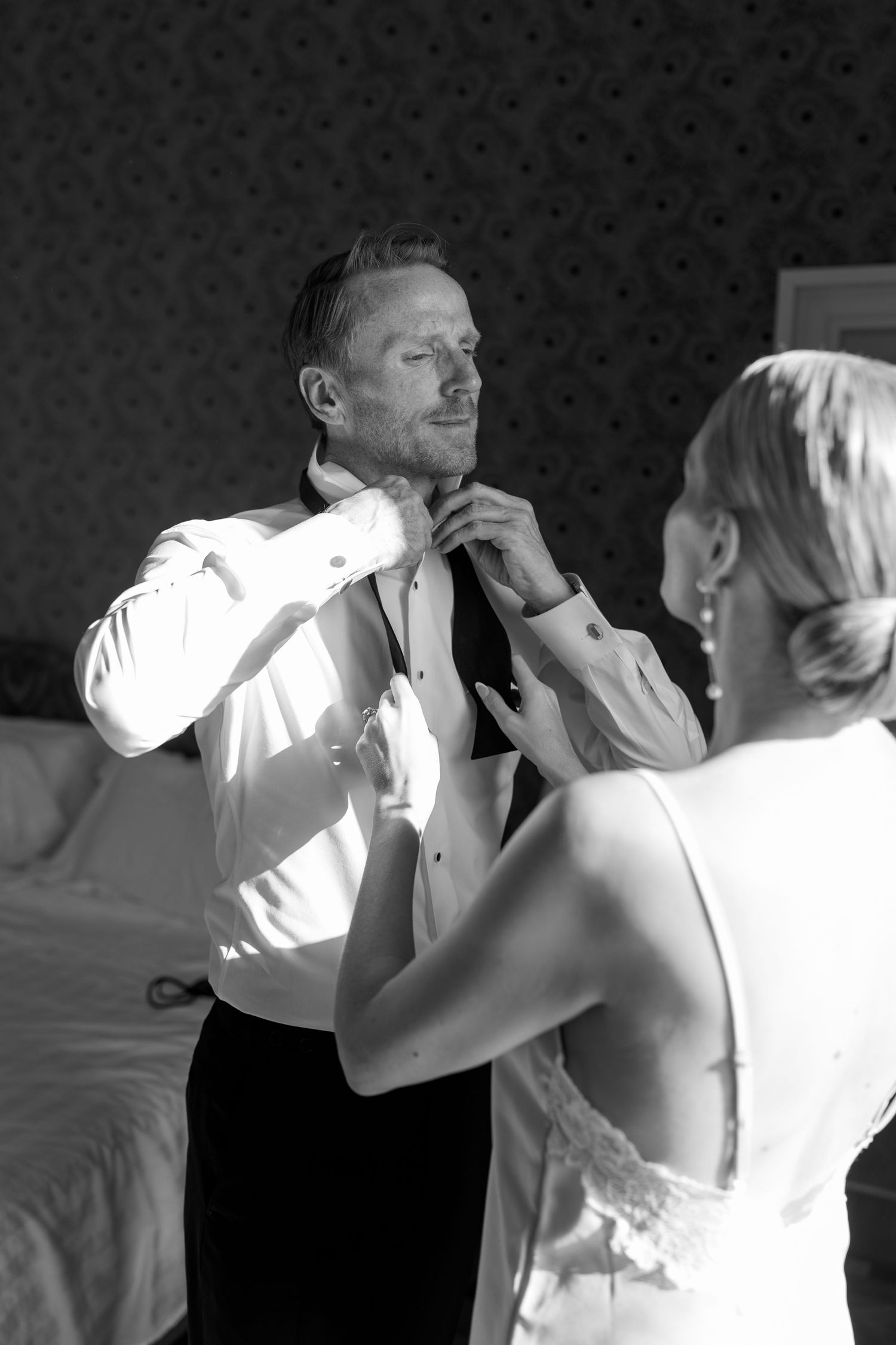 Black and white photo of bride helping groom adjust his tie in a hotel room.