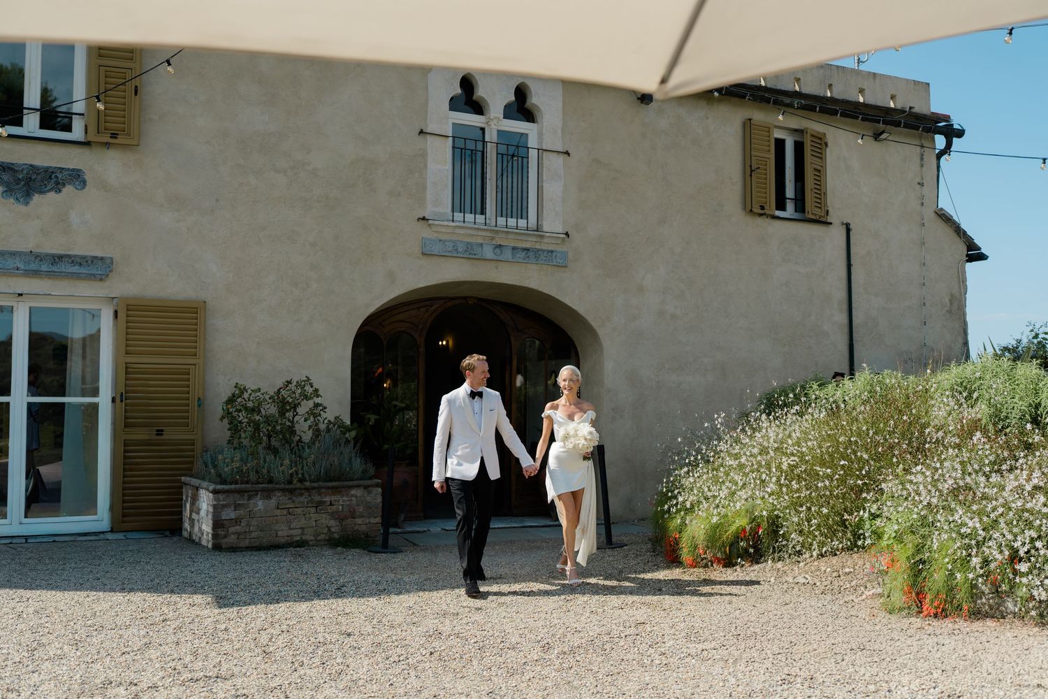 Bride and groom walk hand in hand from rustic stone villa on sunny wedding day.