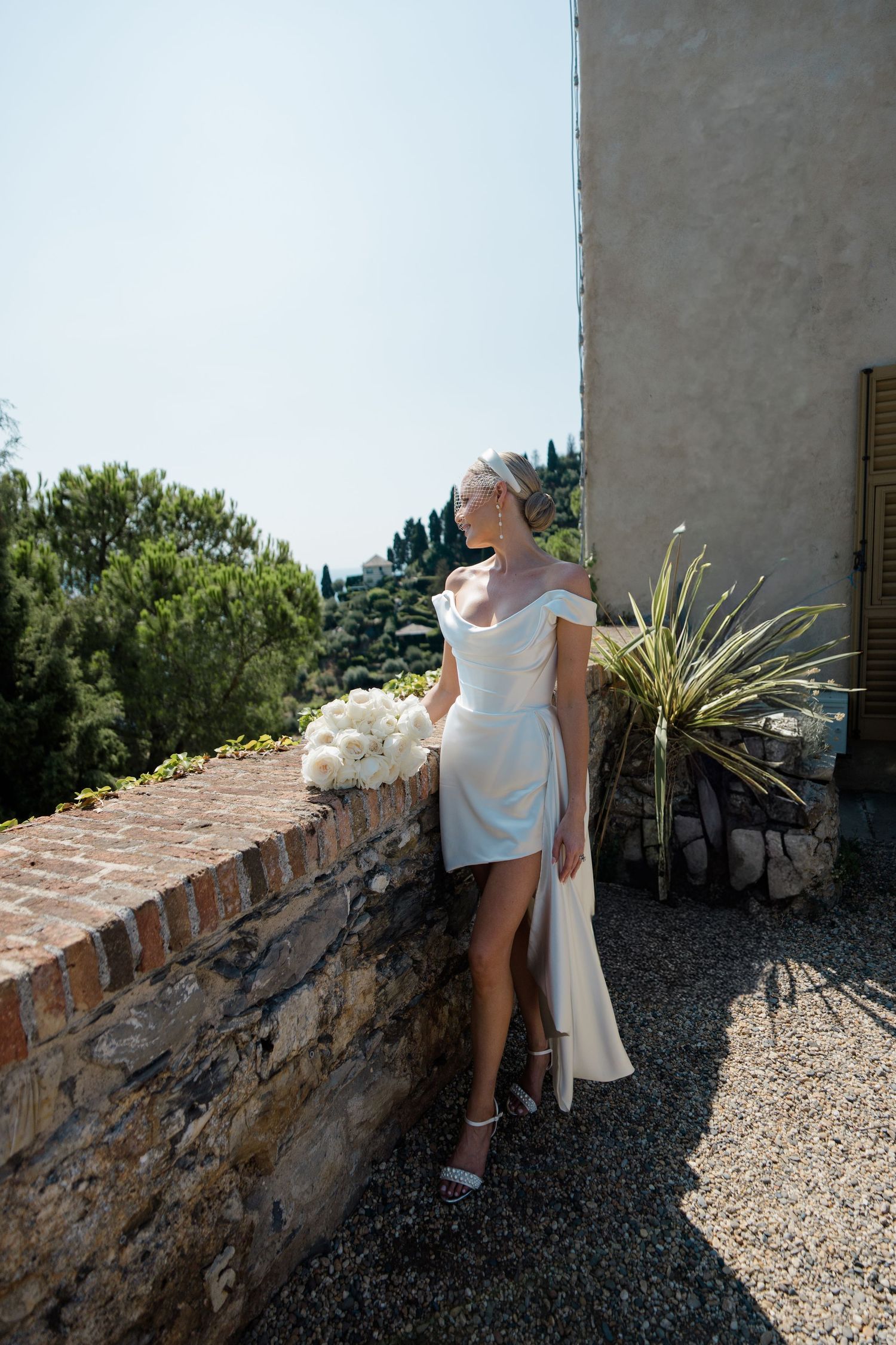 Elegant bride in white off-shoulder mini dress holding bouquet beside rustic brick wall outdoors.
